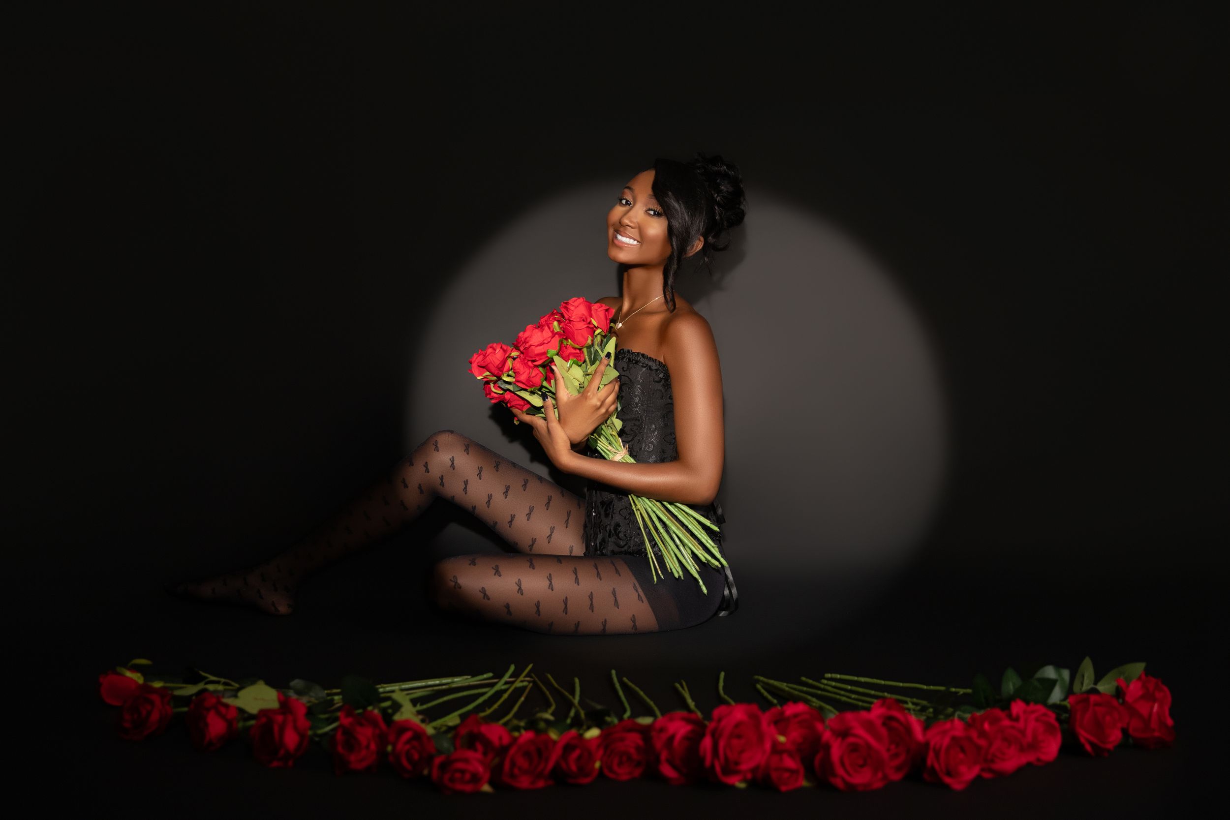 Woman in black lace outfit holds red roses bouquet, sitting among roses on floor in dramatic spotlight.