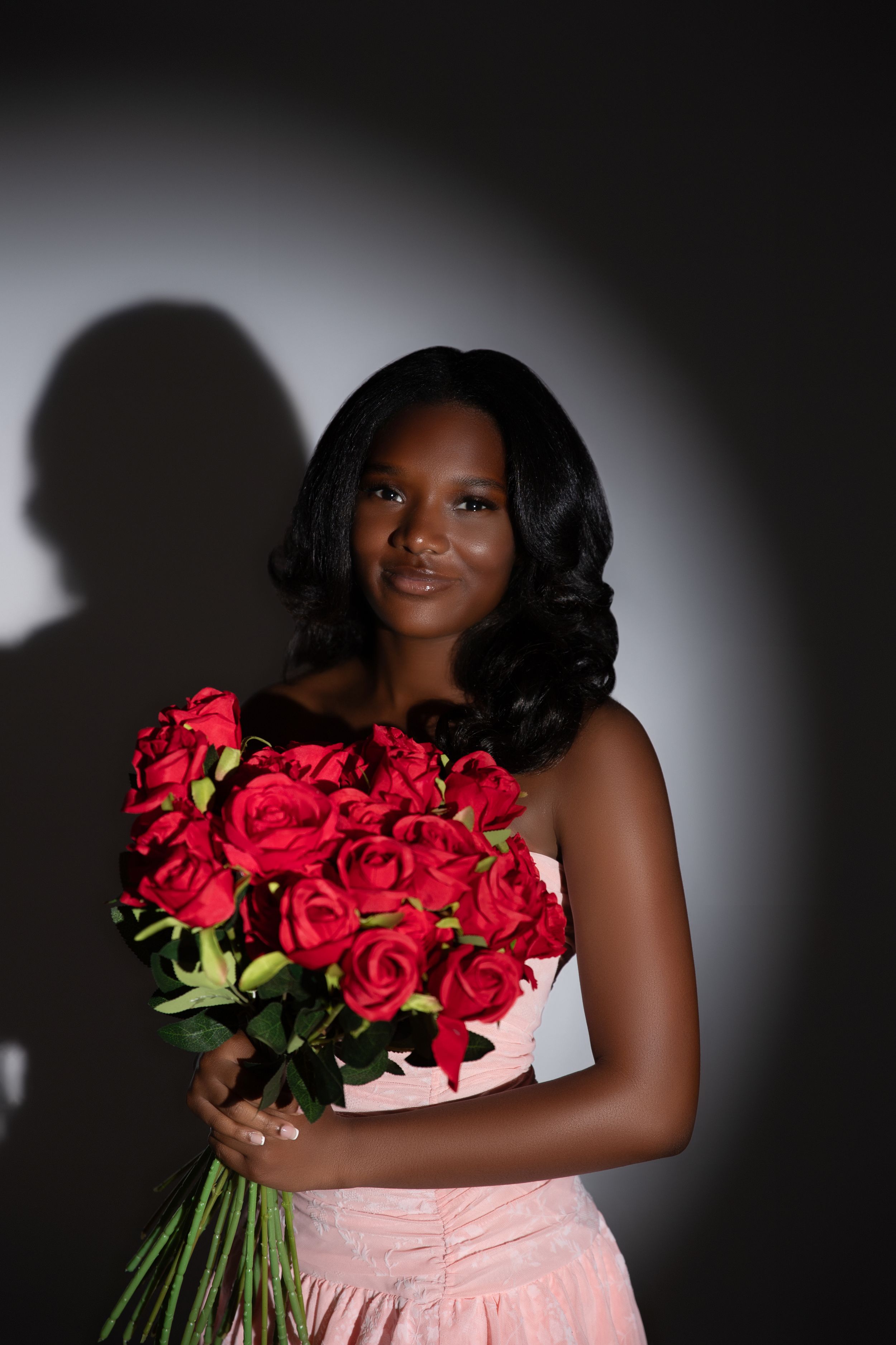 Young Black woman in pink dress holding large bouquet of red roses, posing elegantly against dark studio background.