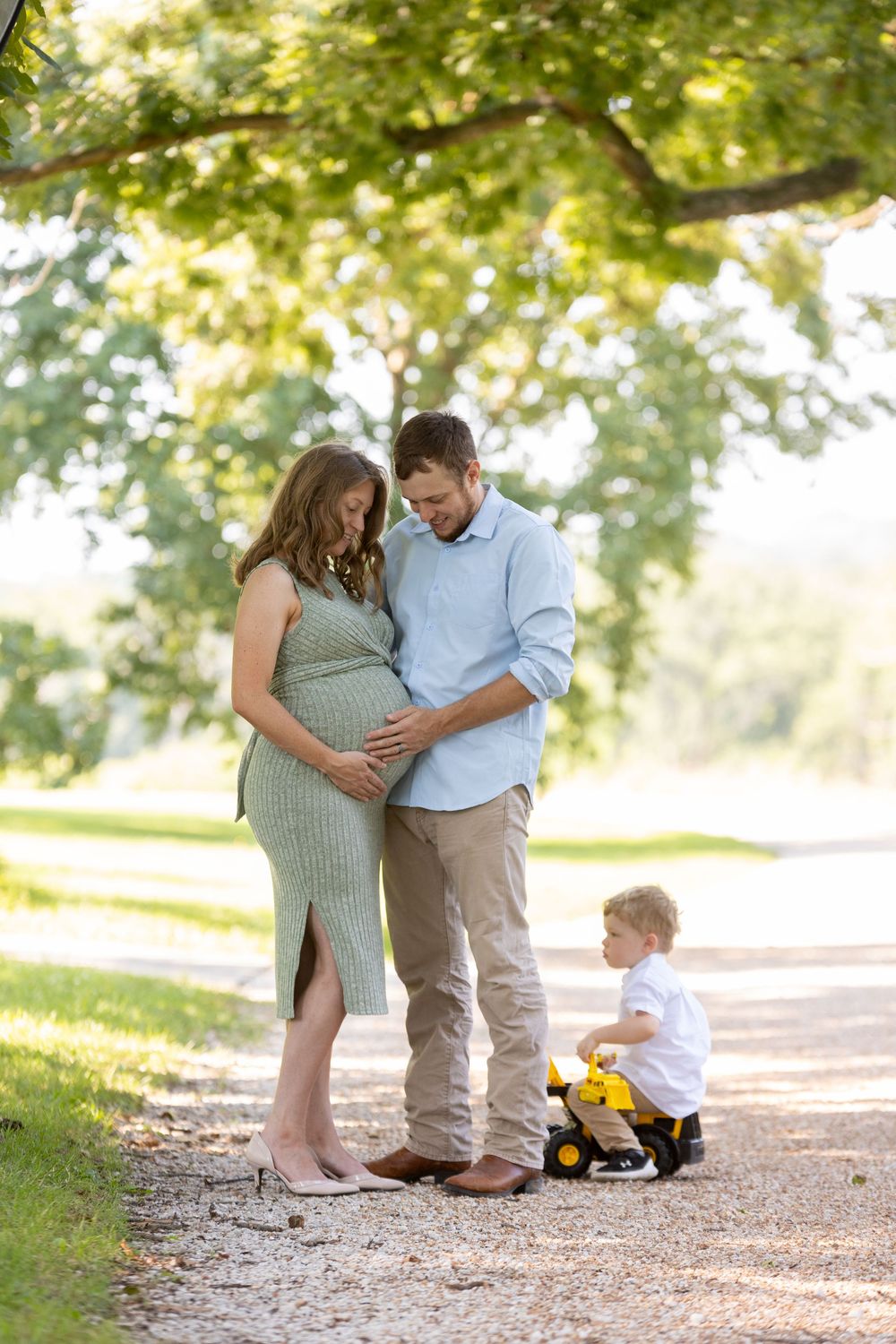 A family shares a tender moment on a tree-lined path as a young child plays with a toy in the background.