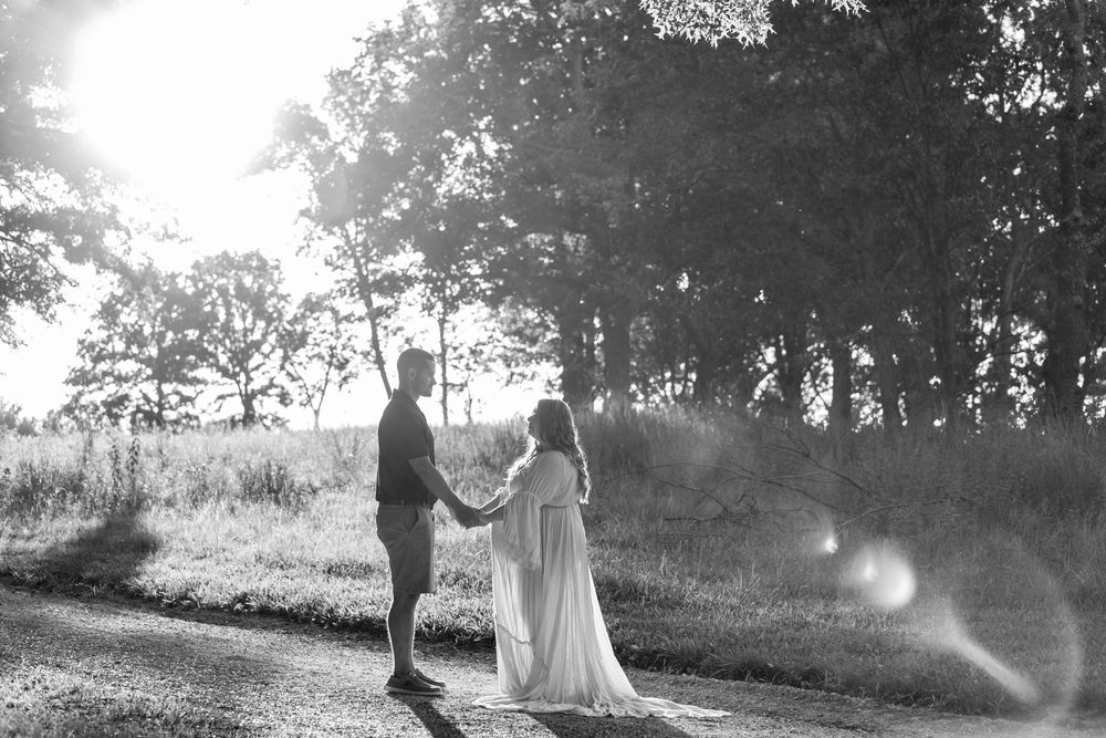 A couple stands together backlit by sunlight in a meadow while wearing elegant wedding attire during golden hour.
