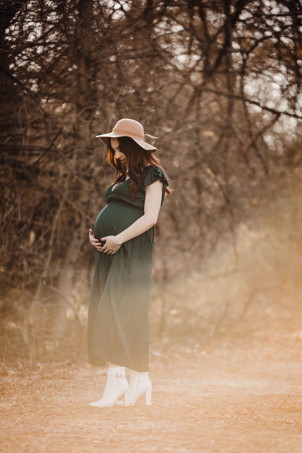 A pregnant woman in a green dress and floppy hat stands in a dreamy autumn setting.