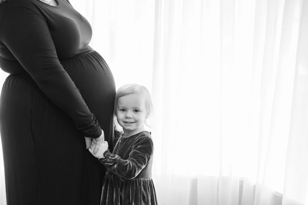 Black and white silhouette of child in velvet dress next to pregnant woman by sheer window.