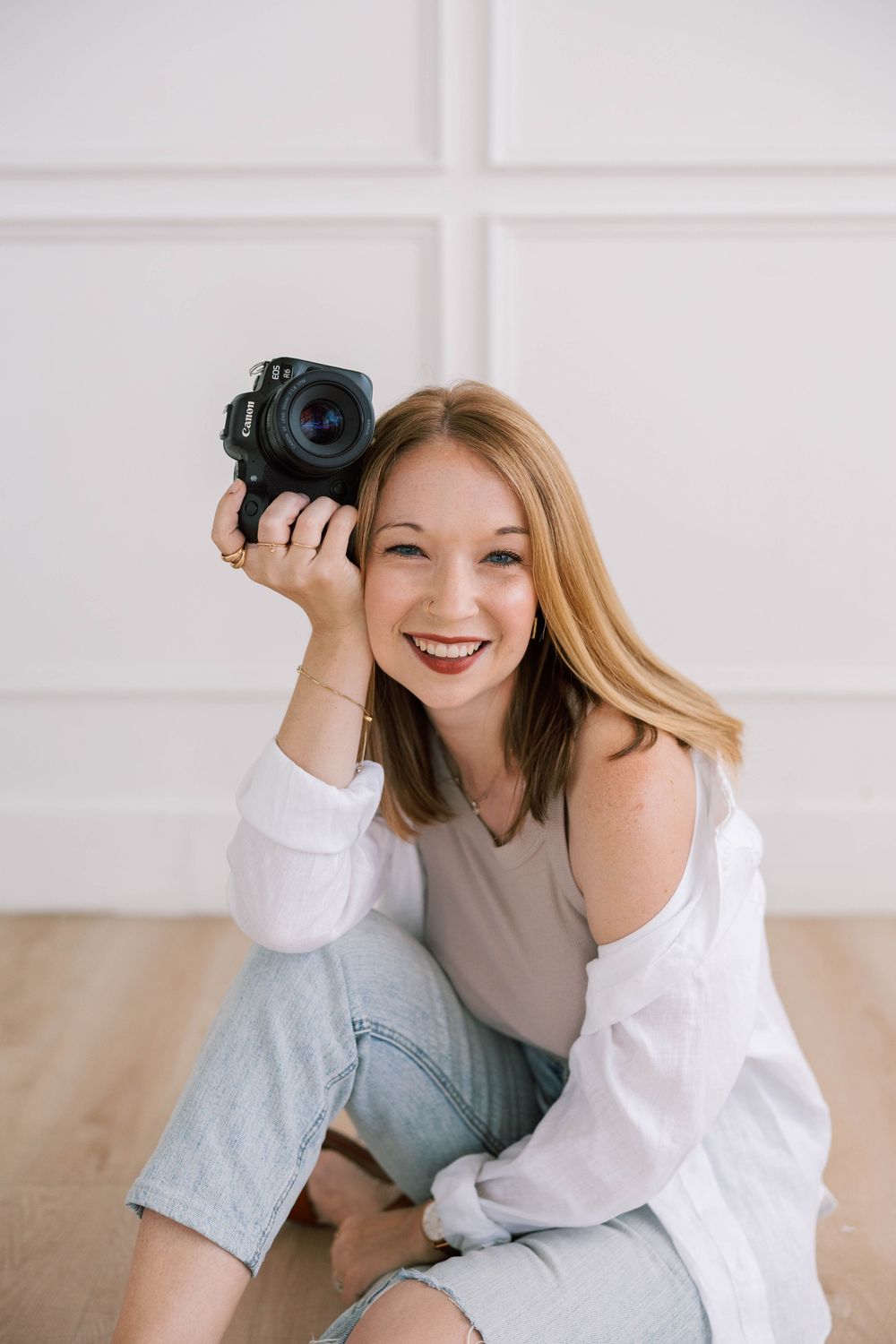 Smiling woman photographer sitting on floor holding black camera, wearing white shirt and jeans in bright white studio in Austin, Texas