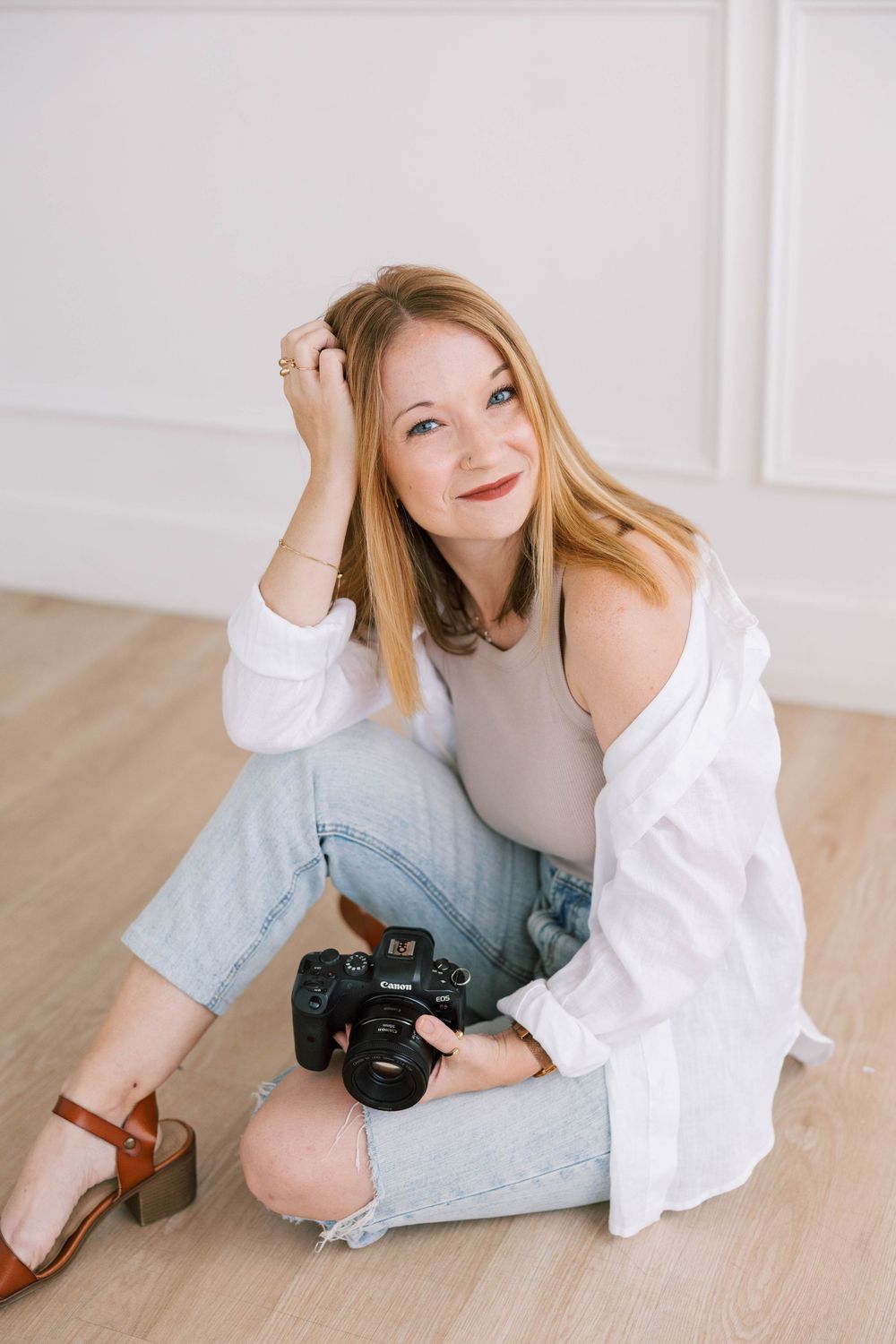 Austin Woman photographer sitting on floor holding DSLR camera, wearing white shirt and jeans, smiling in bright studio.
