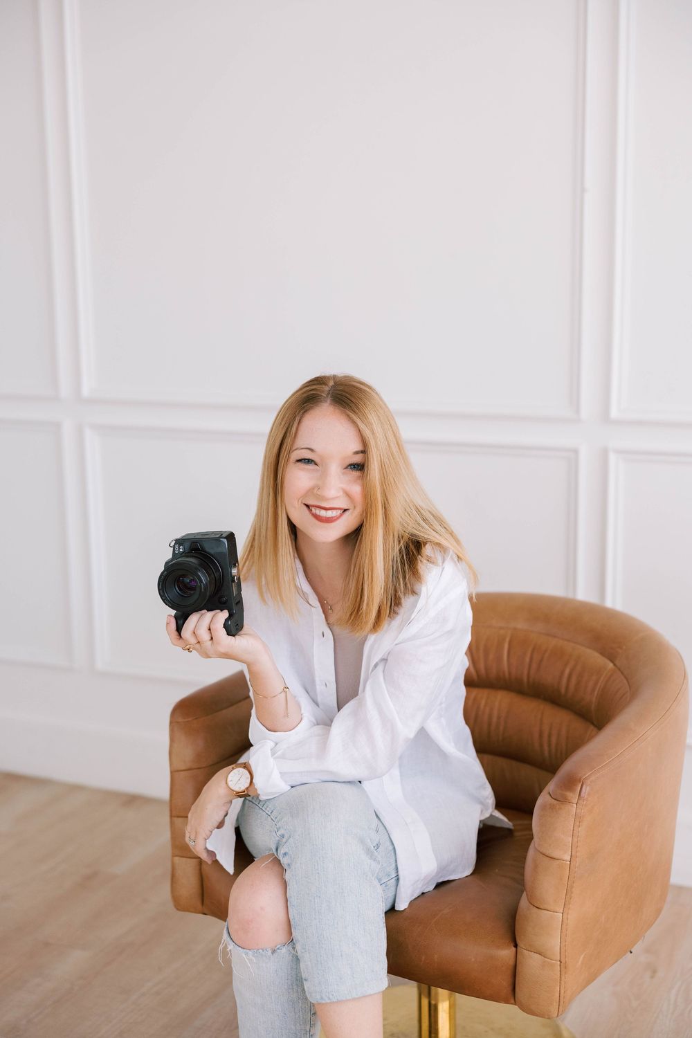 Smiling blonde woman sitting in brown leather chair holding a black camera, wearing white shirt and ripped jeans in Austin, Texas