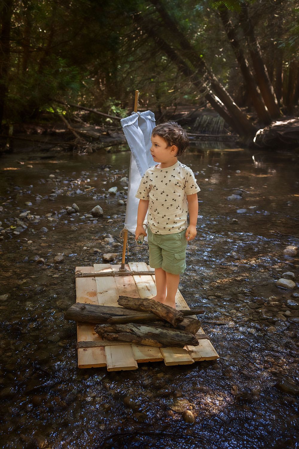 A child explores a rocky creek bed while wearing a light shirt and olive green shorts in a wooded setting.