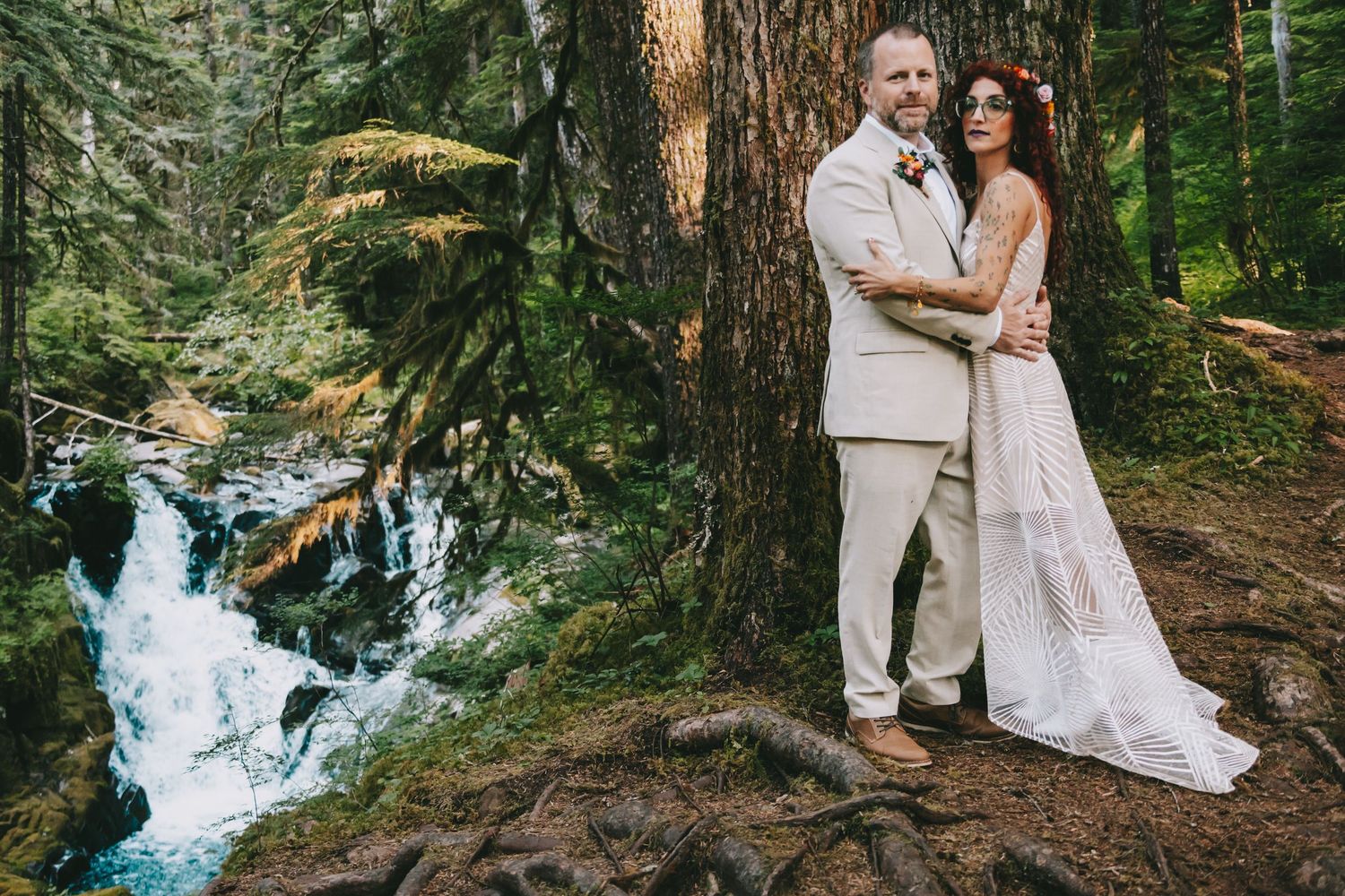 Bride and groom embrace in front of a tree with lots of roots. Waterfall flows in the background of left side. Olympic National Park