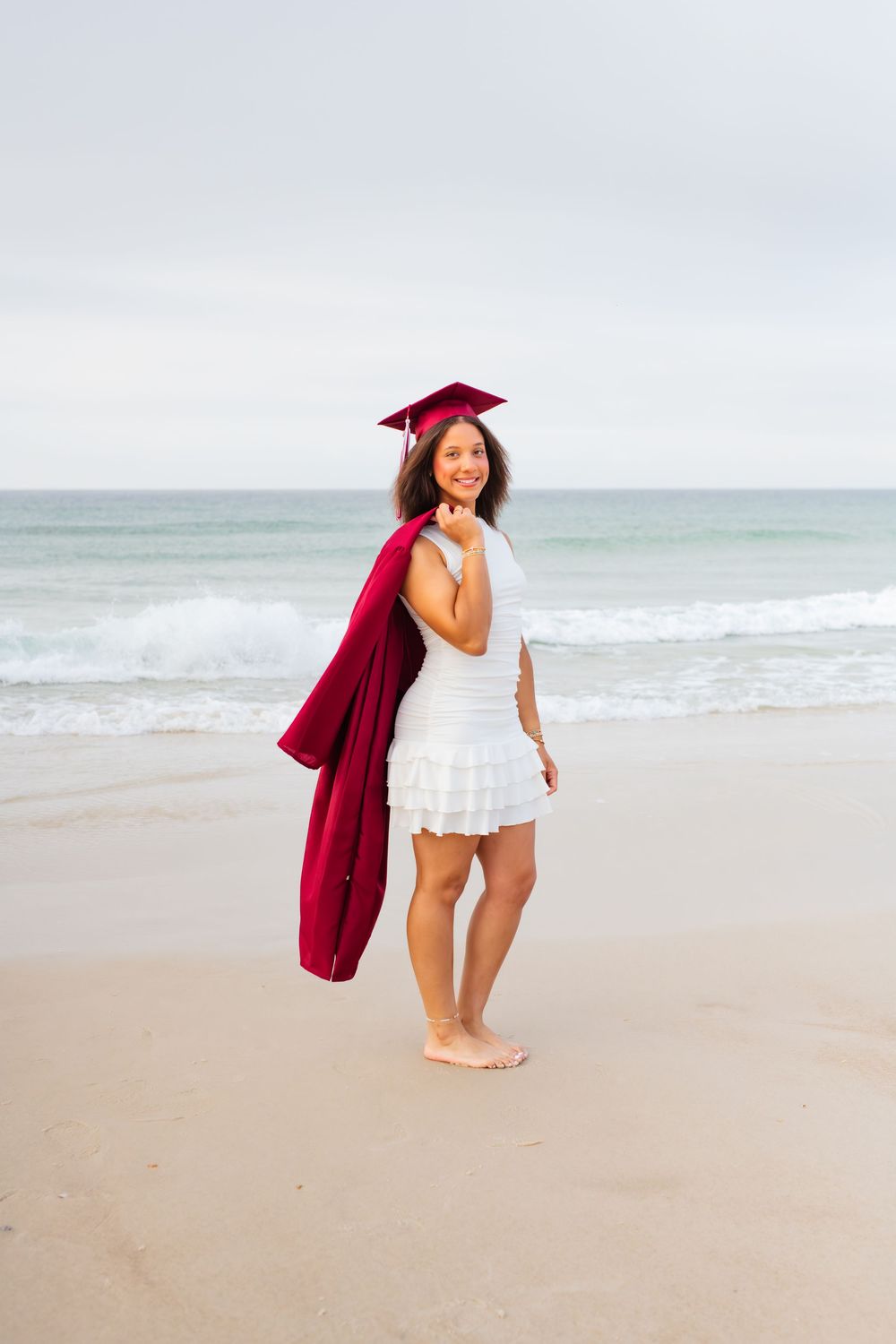 Smiling graduate in white dress and maroon cap and gown poses barefoot on sandy beach with ocean waves behind her.