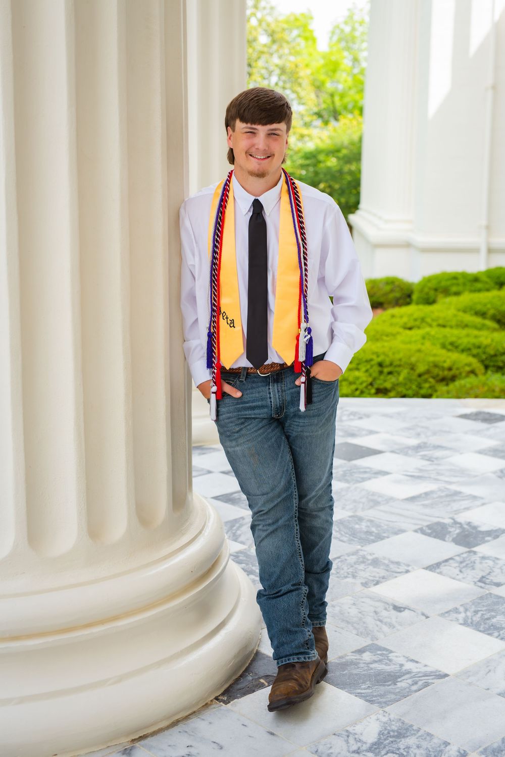 Smiling graduate in white shirt, tie, and colorful stole leans against white column outdoors.