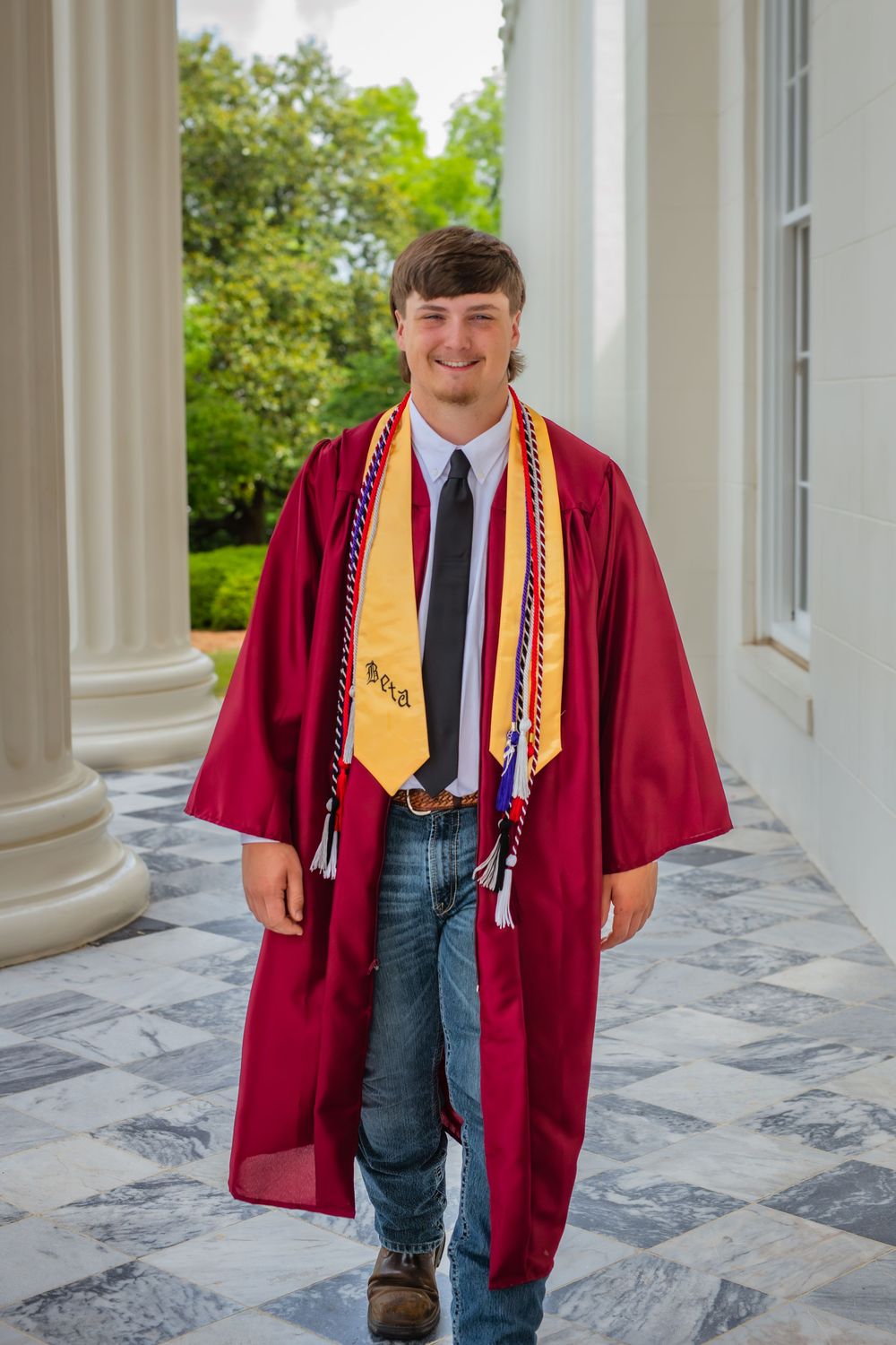 Smiling young man in red graduation gown with gold stole and honor cords poses between white columns.