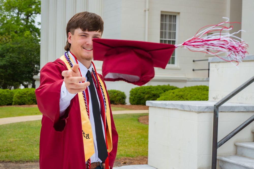 College graduate in red cap and gown tossing tassel outdoors near white building columns on graduation day.