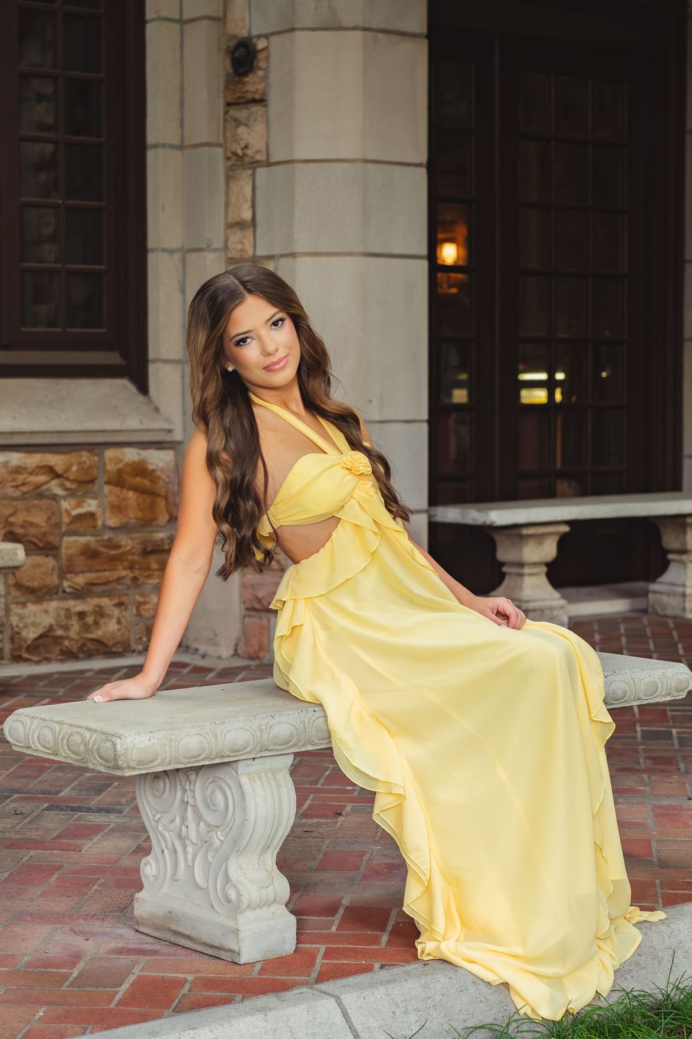Young woman in a yellow ruffled gown sitting on a stone bench outside a grand building.