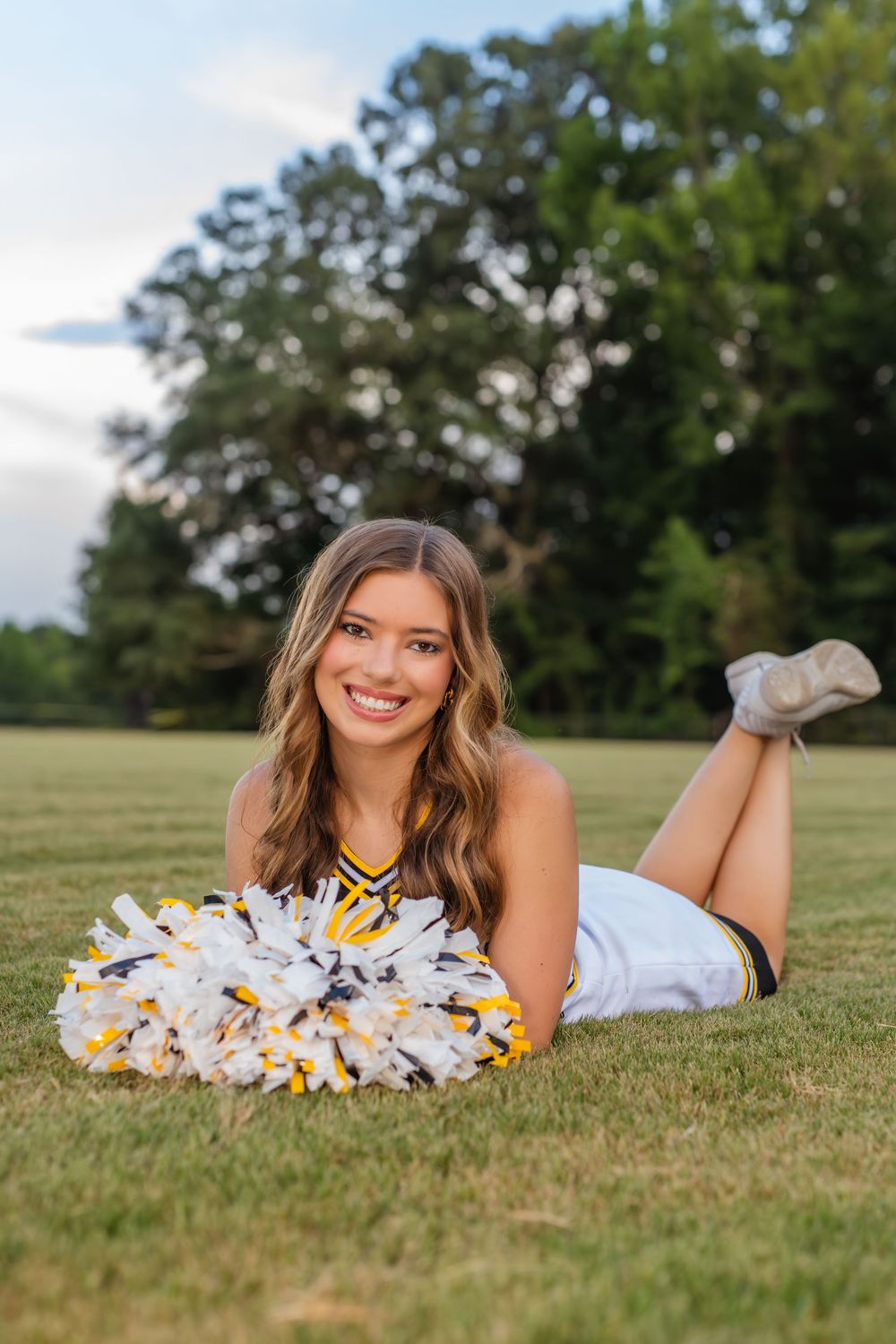 Smiling cheerleader in white and gold uniform lying on grass field holding pom-poms with trees in background.