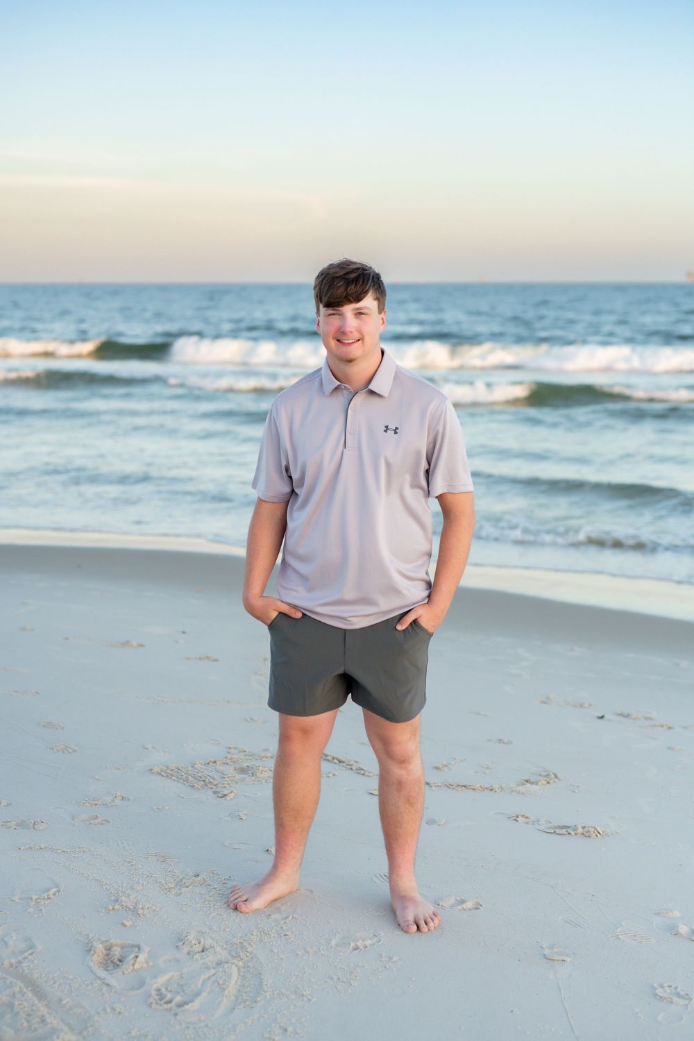 A casual beach scene showing someone wearing a light gray t-shirt and dark shorts while standing on sandy shoreline at sunset.