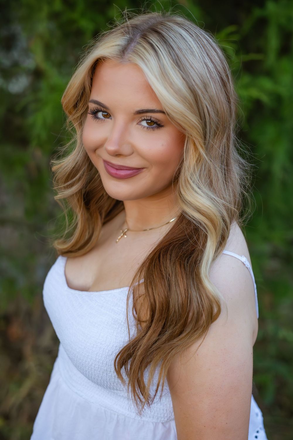 Portrait of someone with wavy blonde hair wearing a white sleeveless top against a blurred green outdoor background.