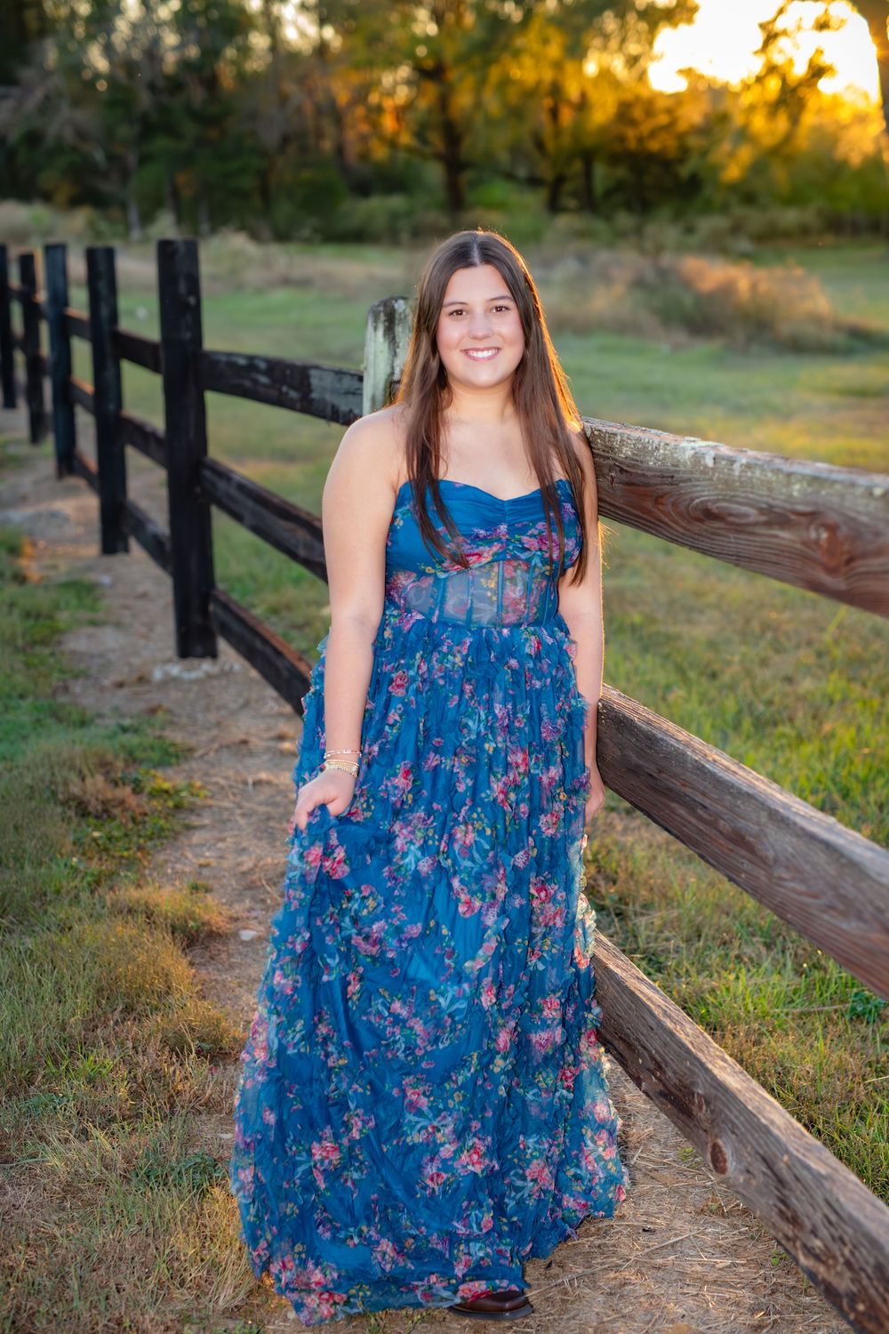A woman in a blue floral dress poses outdoors near a rustic wooden fence during golden hour.