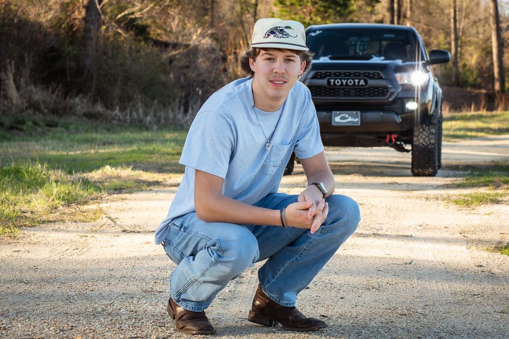 Young man in gray shirt and jeans crouching on gravel path in front of a black Toyota truck outdoors.