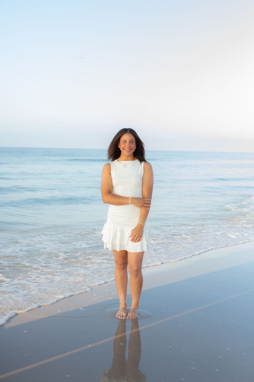 A smiling woman in a white dress stands barefoot on a sandy beach with gentle ocean waves behind her.