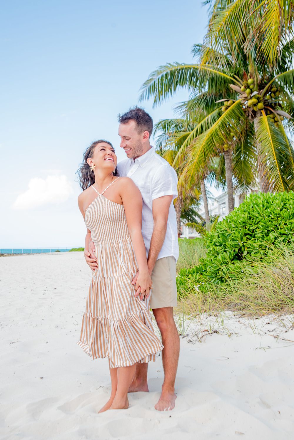 Smiling couple holding hands on a tropical beach with palm trees, woman in striped dress, man in white shirt.