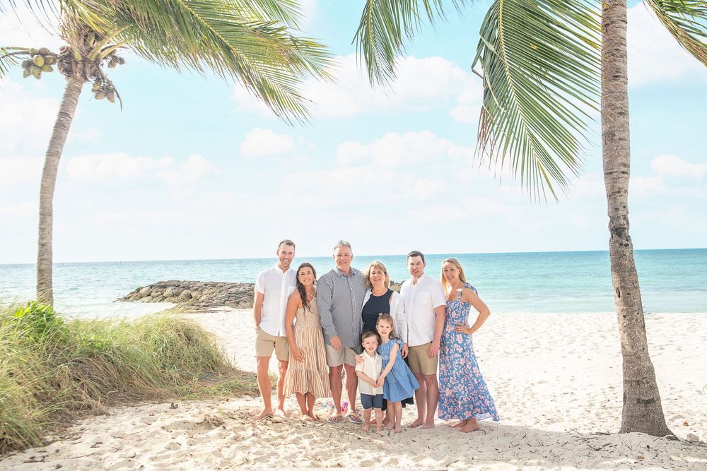 Family of seven posing together on a tropical beach with palm trees and turquoise ocean in background.
