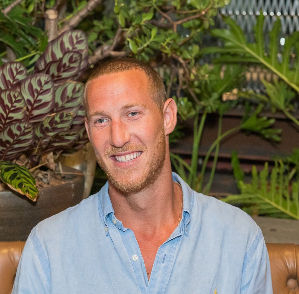 A person in a light blue button-down shirt smiles while seated in front of tropical plants and foliage.