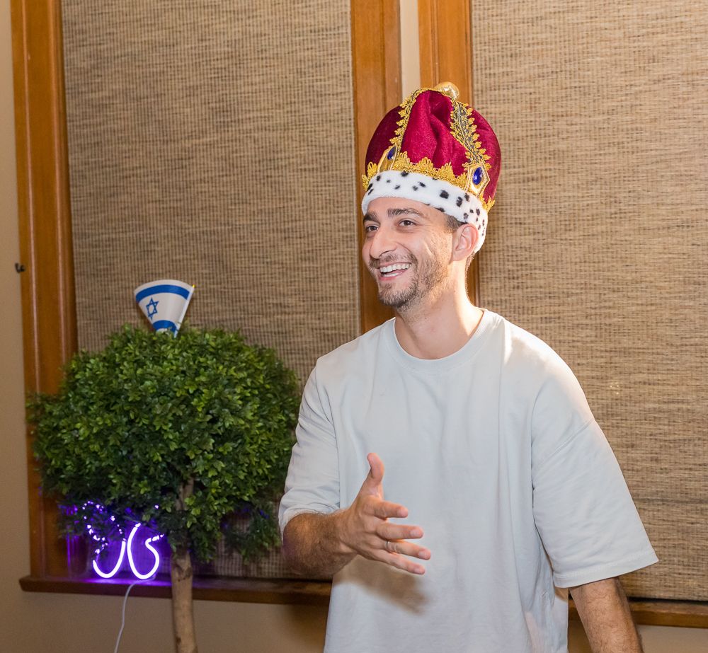 Two people wearing paper crowns smile and gesture while having a conversation in front of a beige wall.