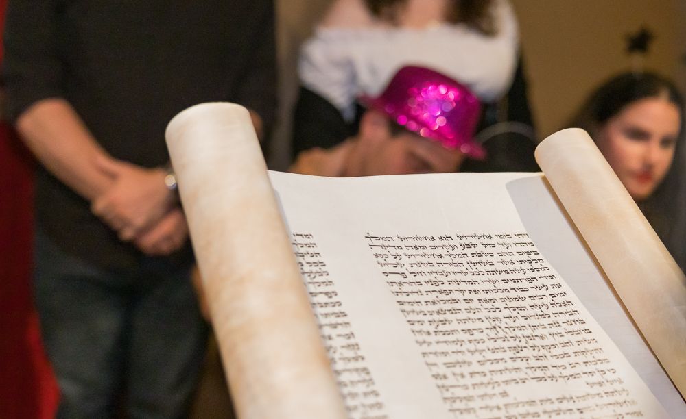 A hand holds open a book against a blurred background with people visible in soft focus.
