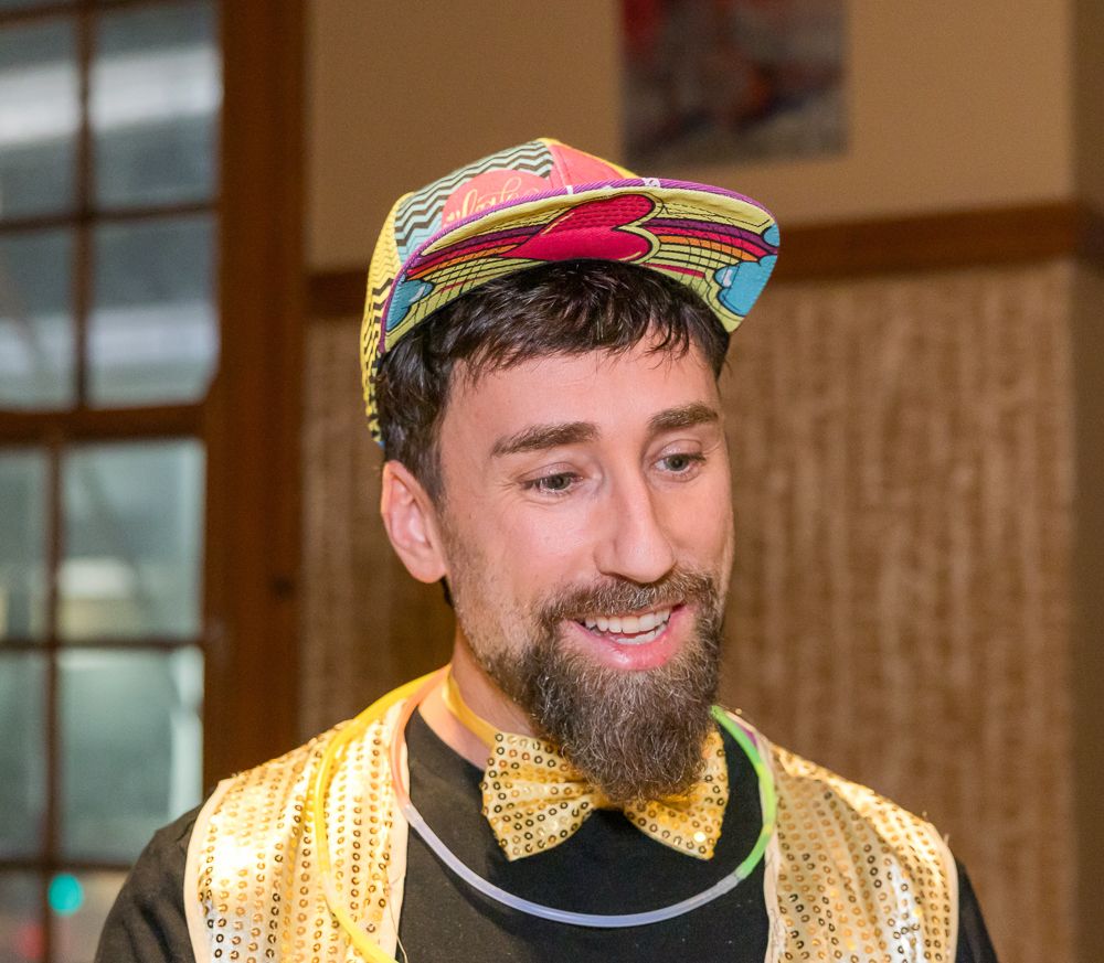 A person wearing a colorful baseball cap smiles while wearing a yellow polka dot bowtie.