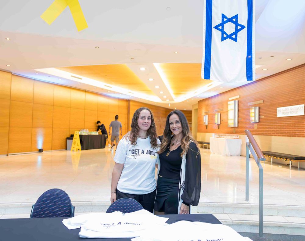 Two people stand in a hallway with an Israeli flag and yellow decorative lighting overhead.