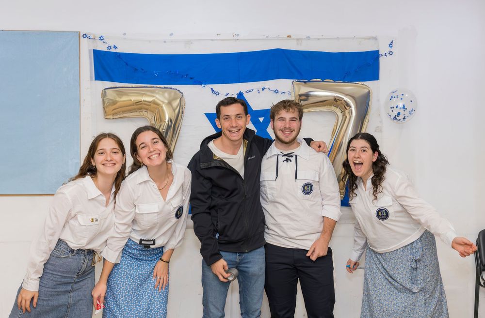 A group celebrates in front of large numbered balloons with a blue banner hanging on the wall behind them.