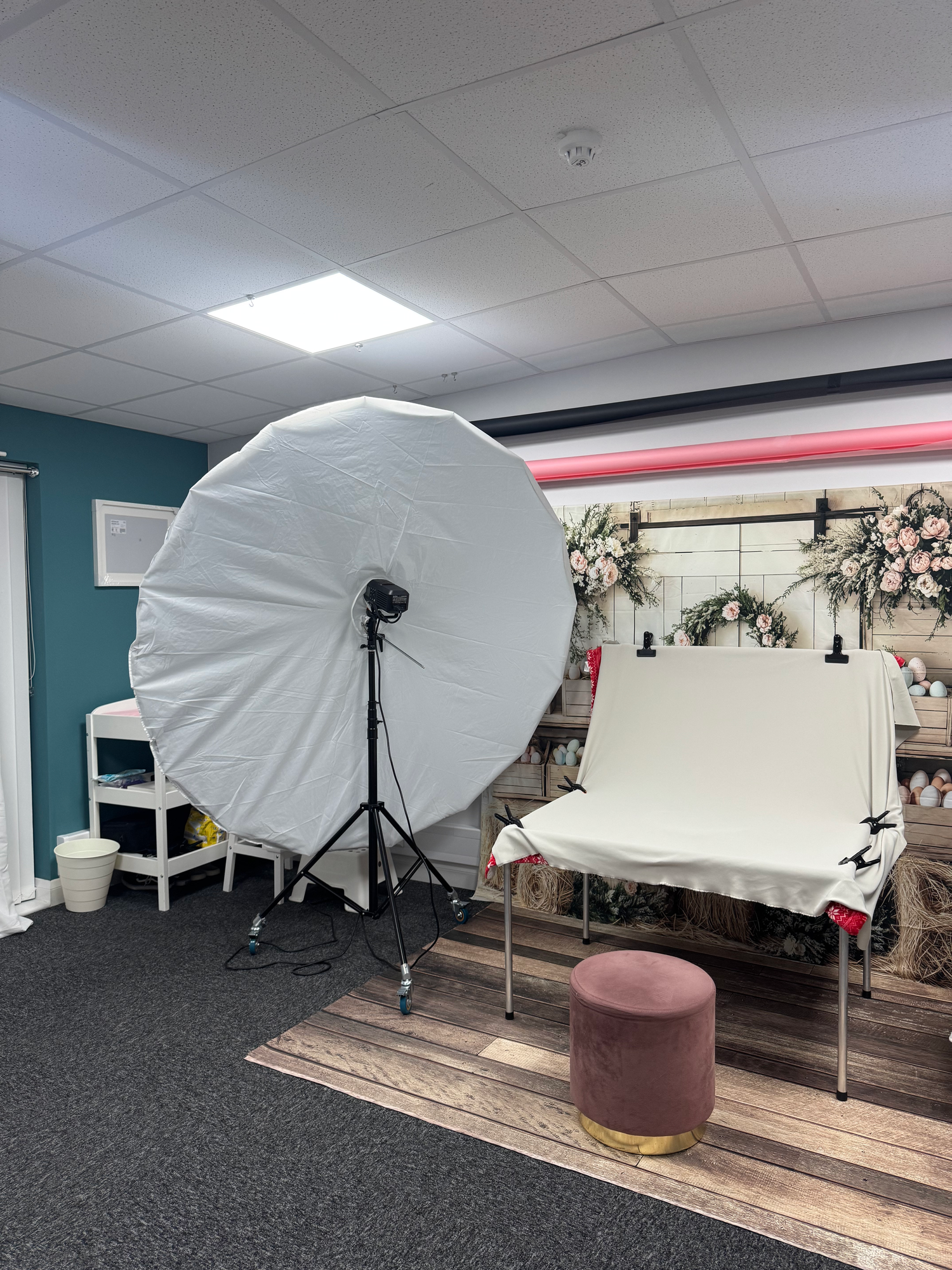 Professional photo studio setup with large white umbrella light, backdrop stand, and pink ottoman on wooden flooring.