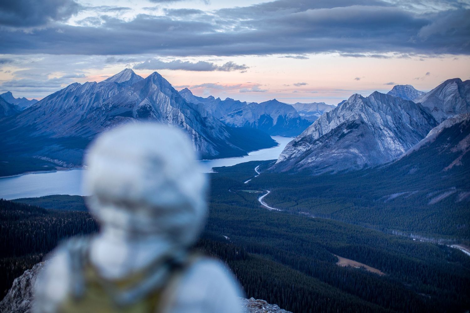 Blurred stone figure overlooking a stunning mountain valley with alpine lakes and snow-capped peaks at dusk.