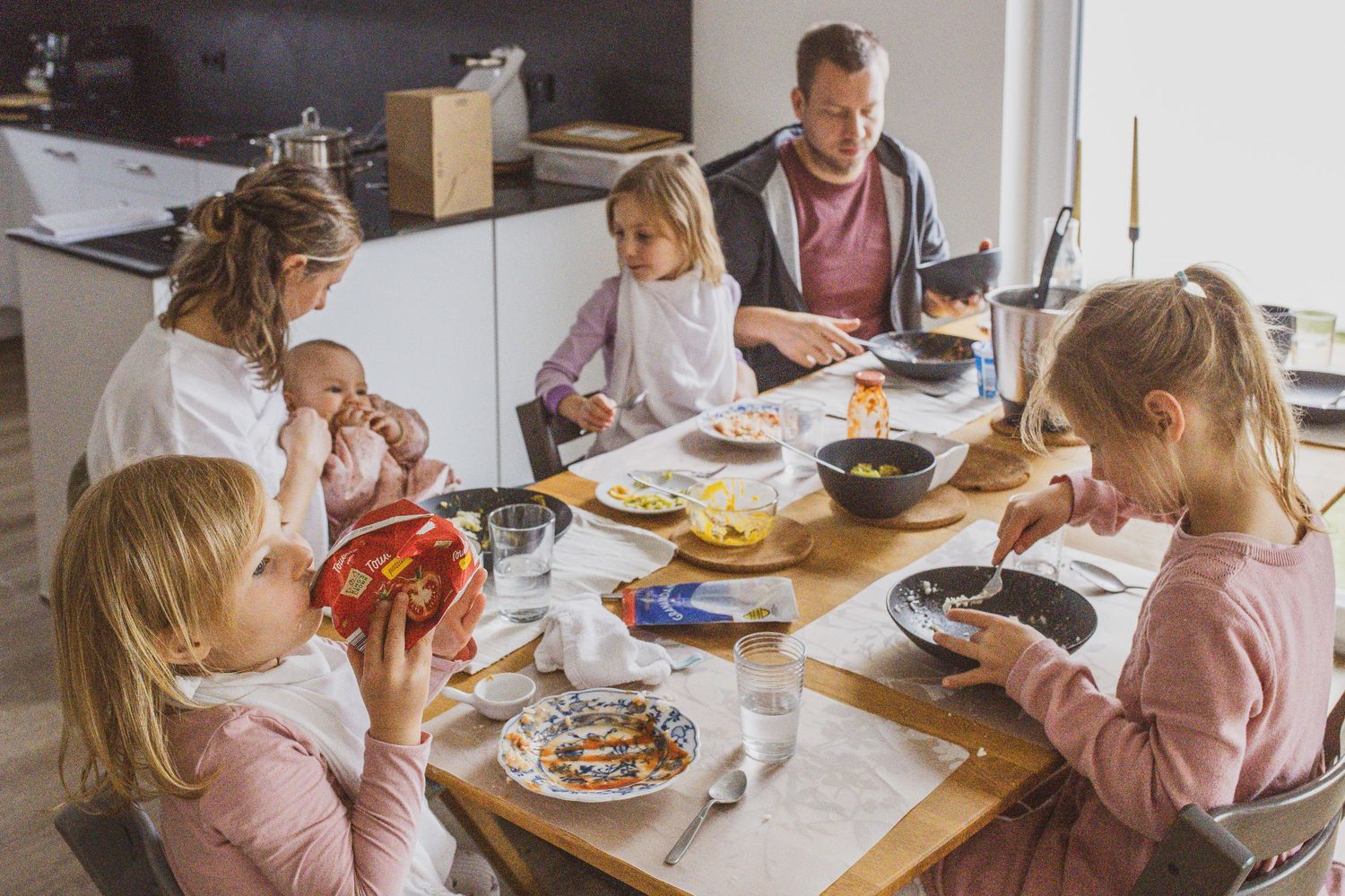 Ungestelltes Familienfoto, im Vordergrund malen die Mutter und zwei Kinder am Tisch mit Wasserfarben, im Hintergrund steht der Vater.
