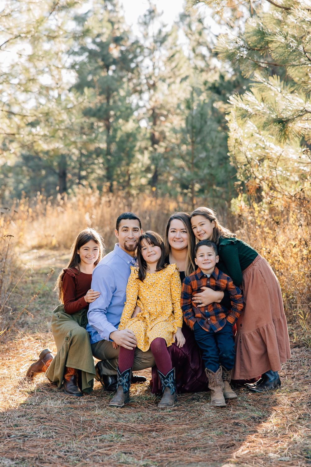 Family group portrait in casual attire sitting together in a sunlit forest setting.