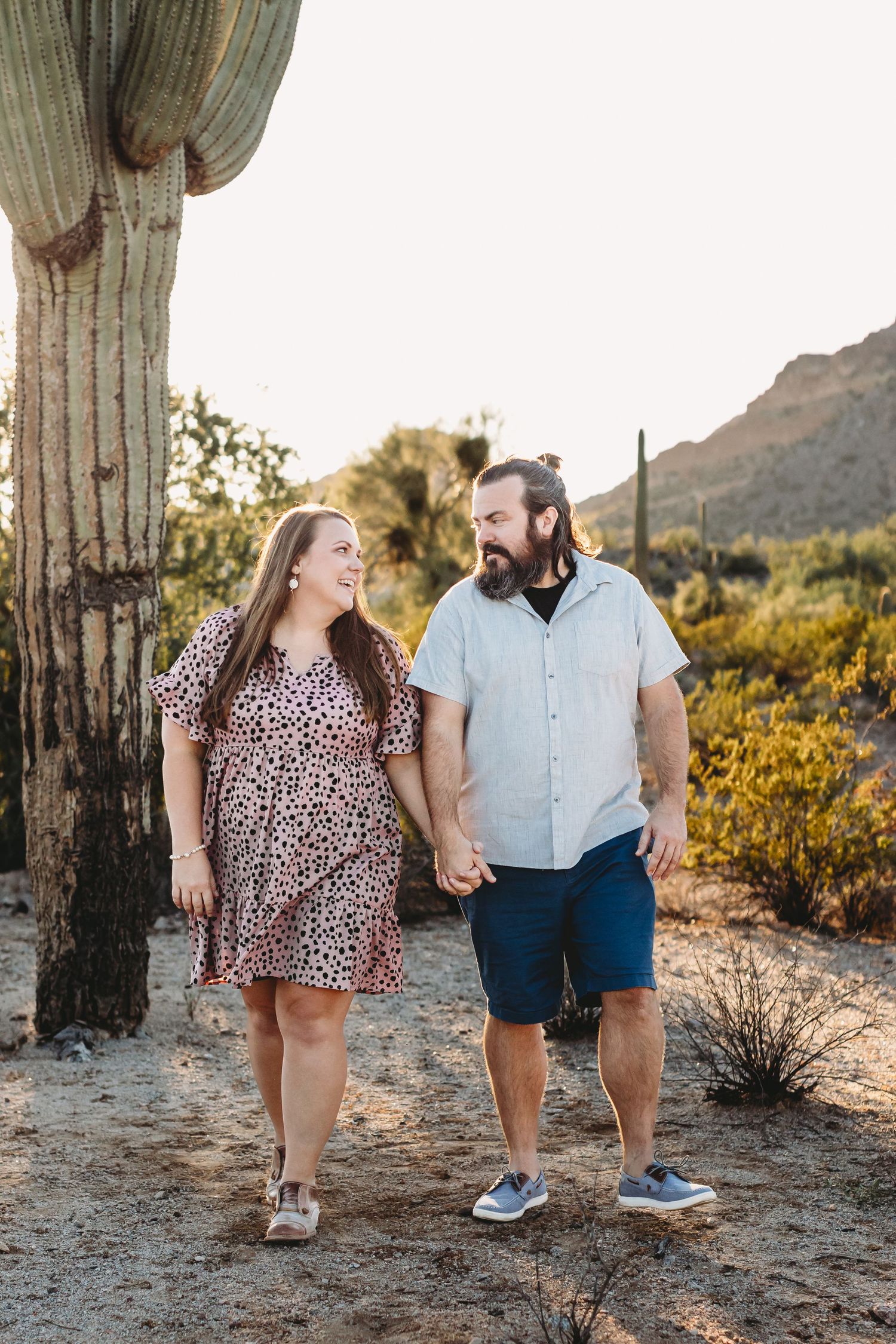 Couple in wedding attire walking along desert path with mountain backdrop.
