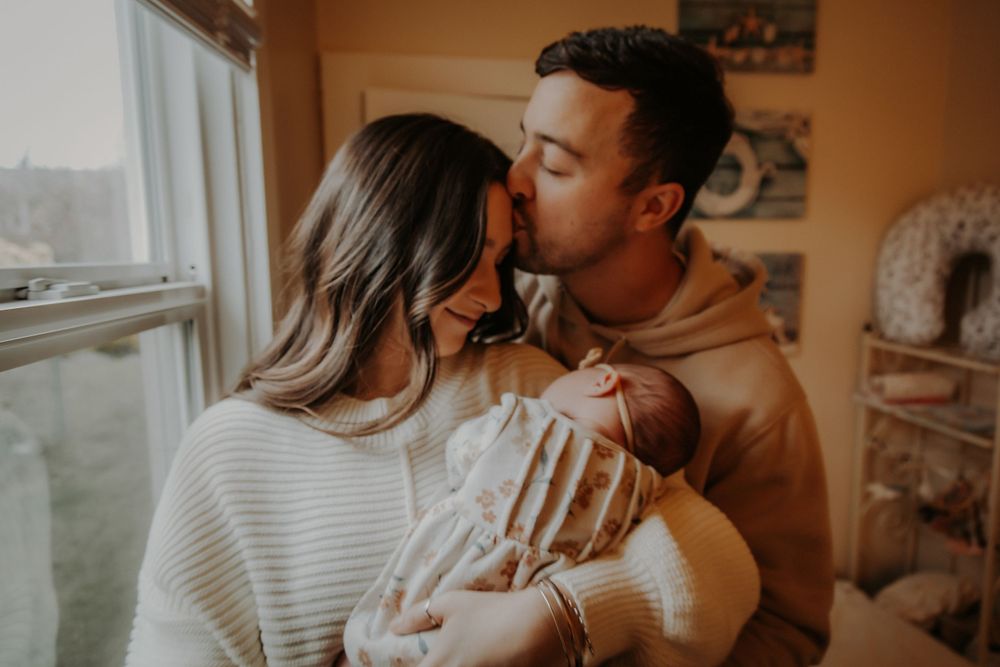 Father kisses mother's forehead as she gently cradles their sleeping newborn baby in a cozy, softly lit nursery.