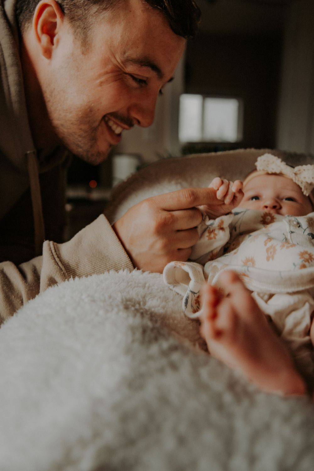 Smiling father gently touches his newborn daughter's hand as she lies bundled in a floral outfit and bow headband.