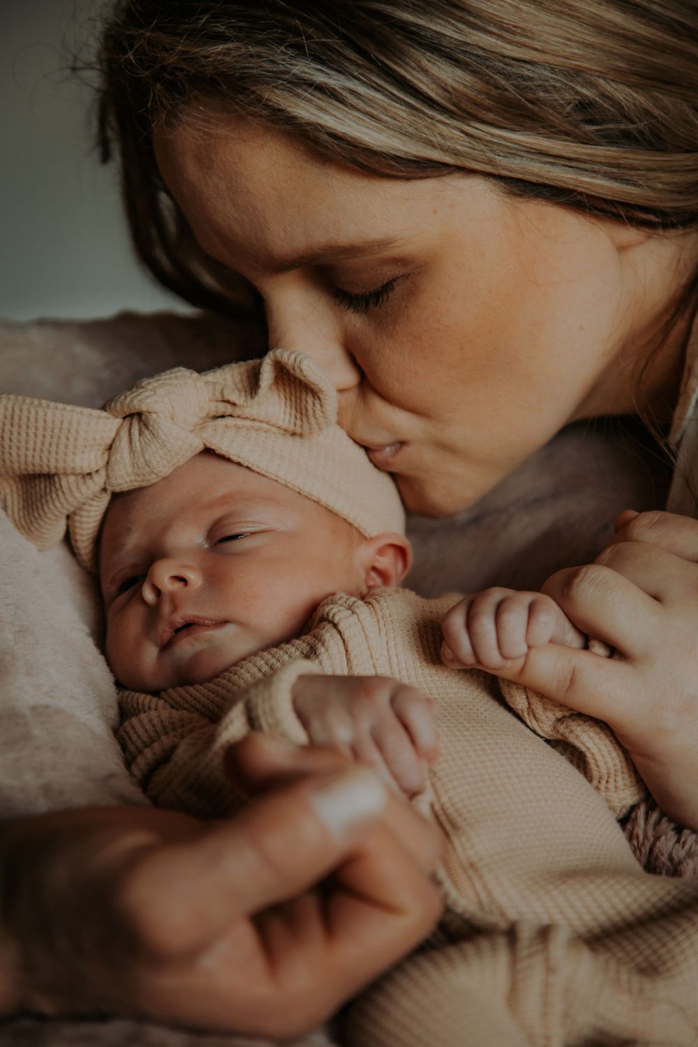 A mother gently kisses the forehead of her sleeping newborn baby wearing a cream knit outfit and matching bow headband.