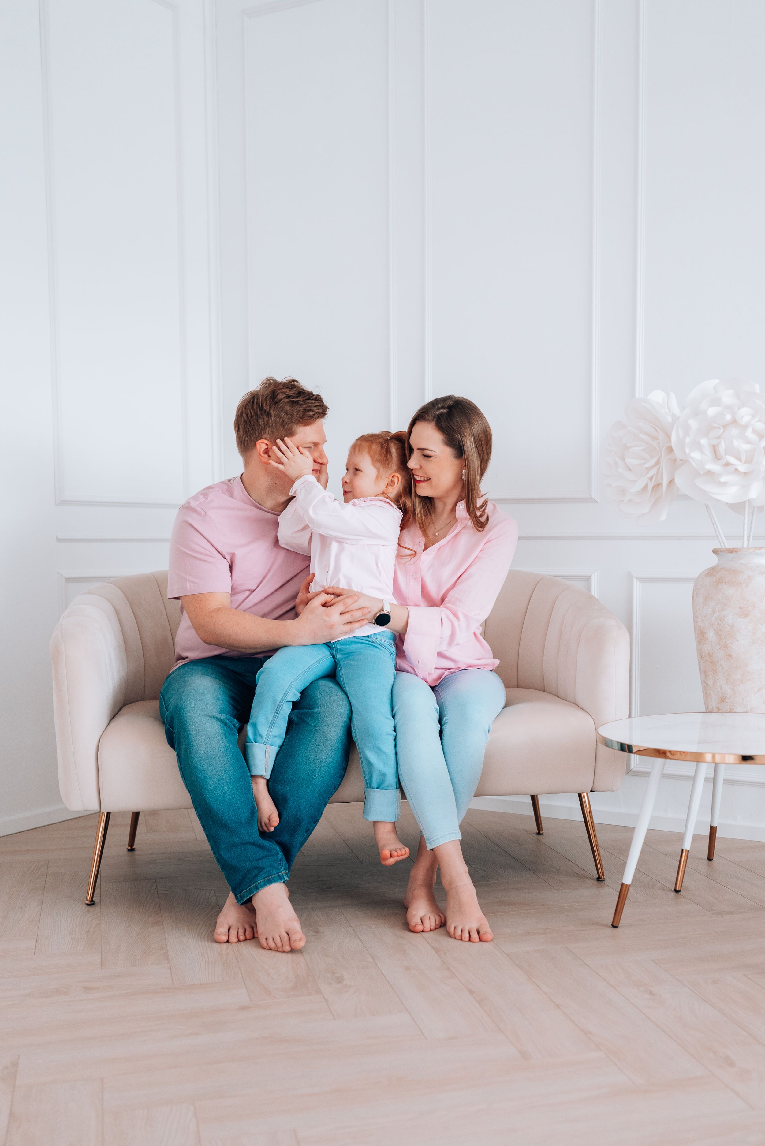 Happy young family with toddler daughter sitting together on beige sofa in bright white living room.