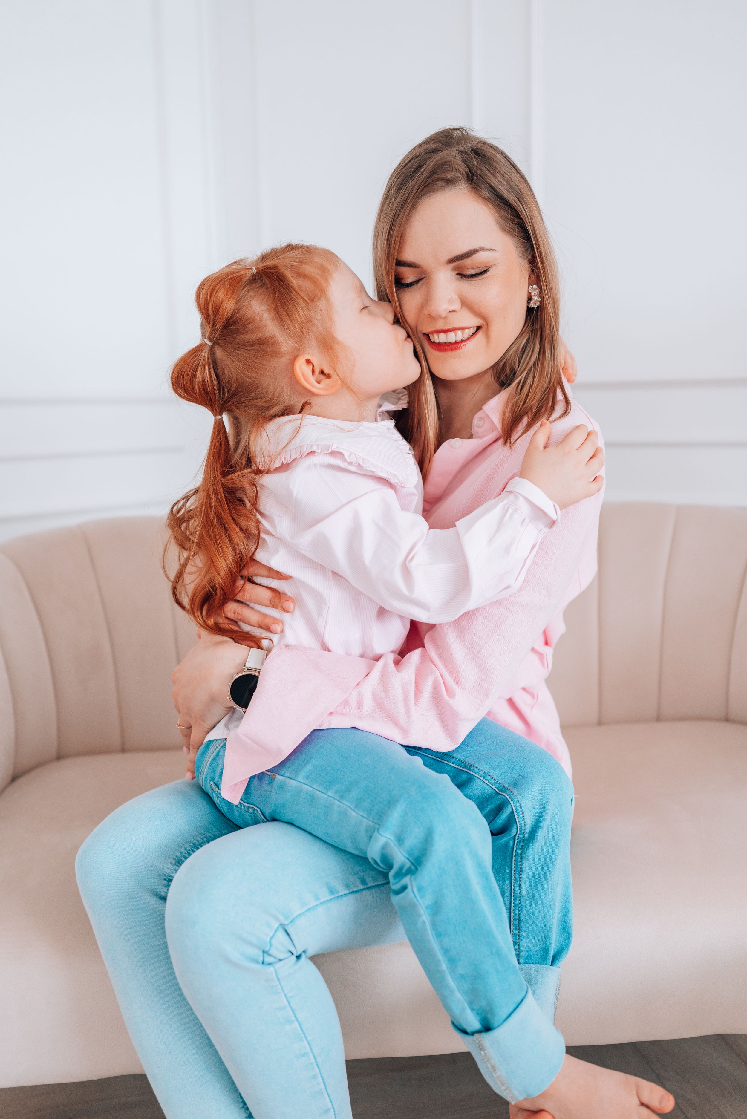 A young redhead girl hugs and kisses her smiling mother sitting on a beige sofa, both wearing pink shirts and blue jeans.