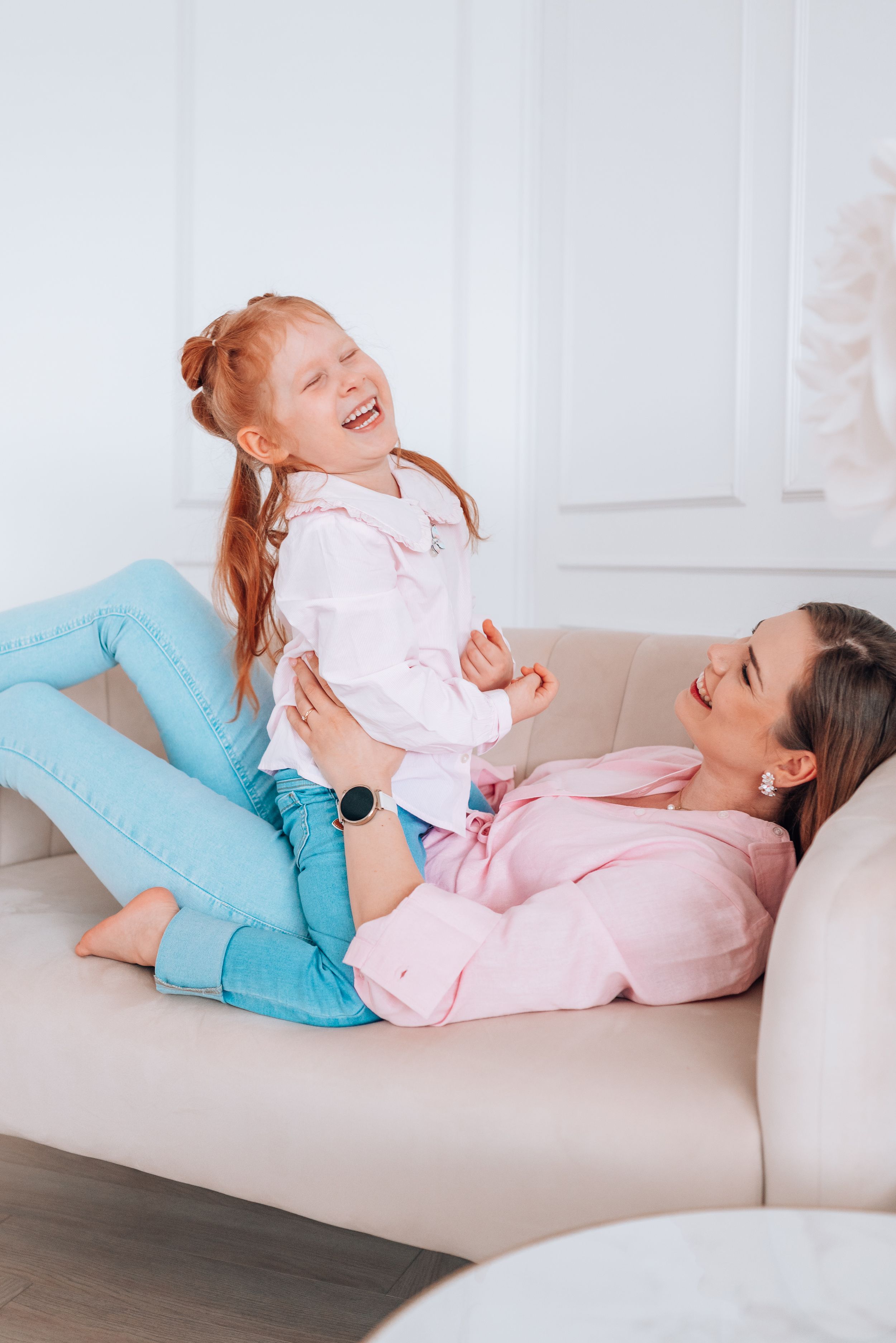 A mother and her red-haired daughter laugh together on a beige sofa, both wearing pink tops and blue jeans.