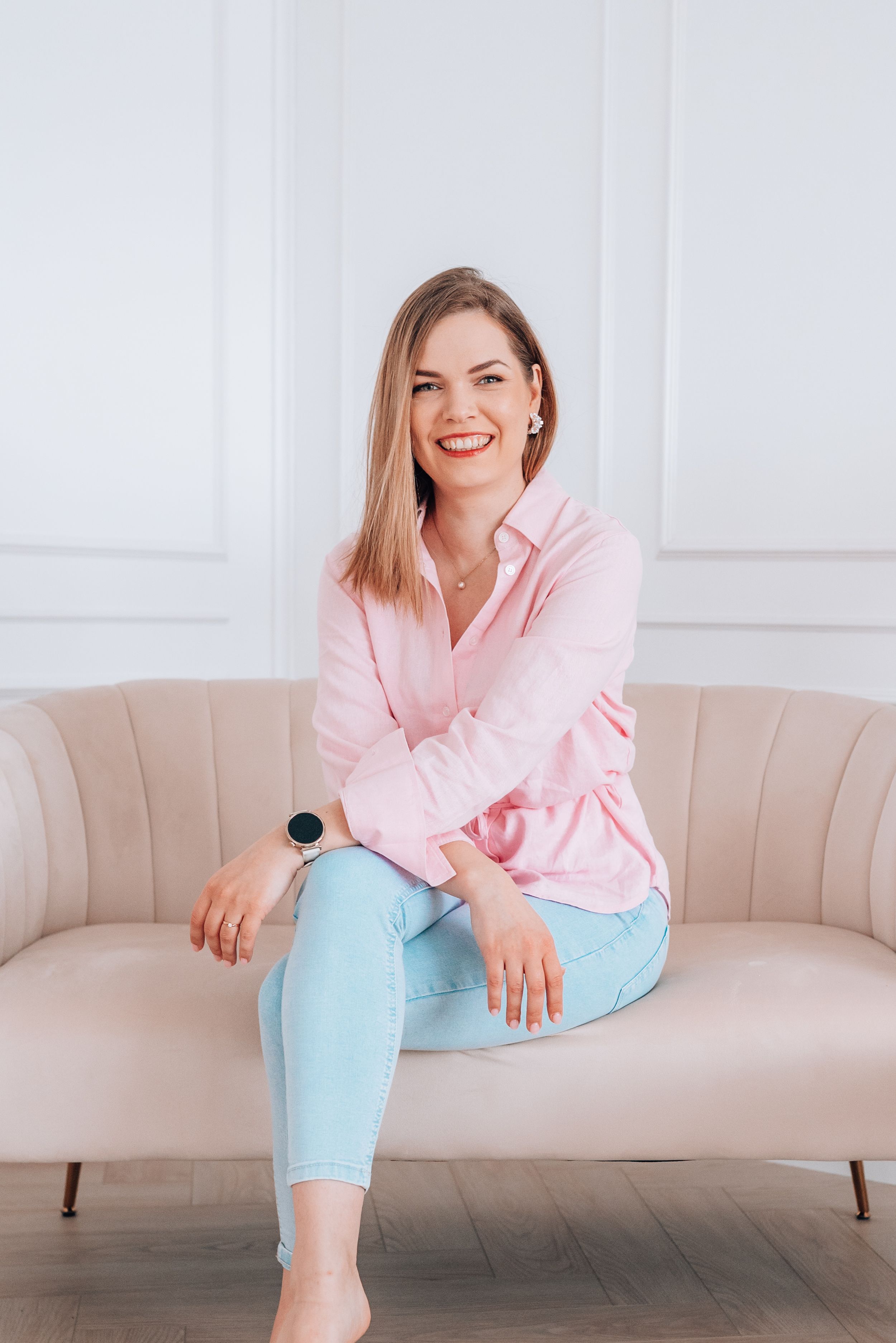 Smiling woman in pink shirt and light blue jeans sitting on a beige sofa in a bright white room.