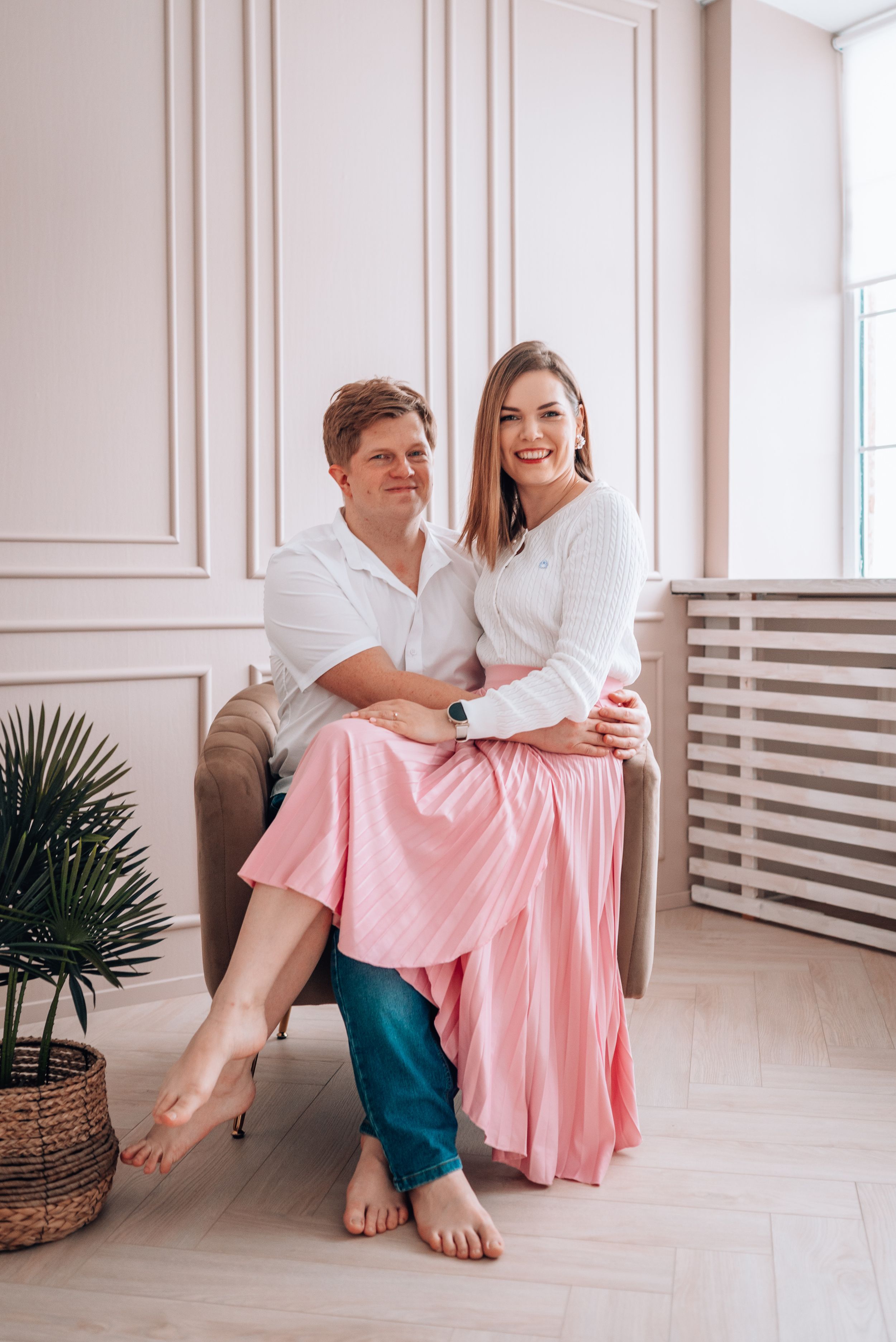 Smiling couple sitting together on chair, woman in pink skirt embracing man in white shirt in elegant room.