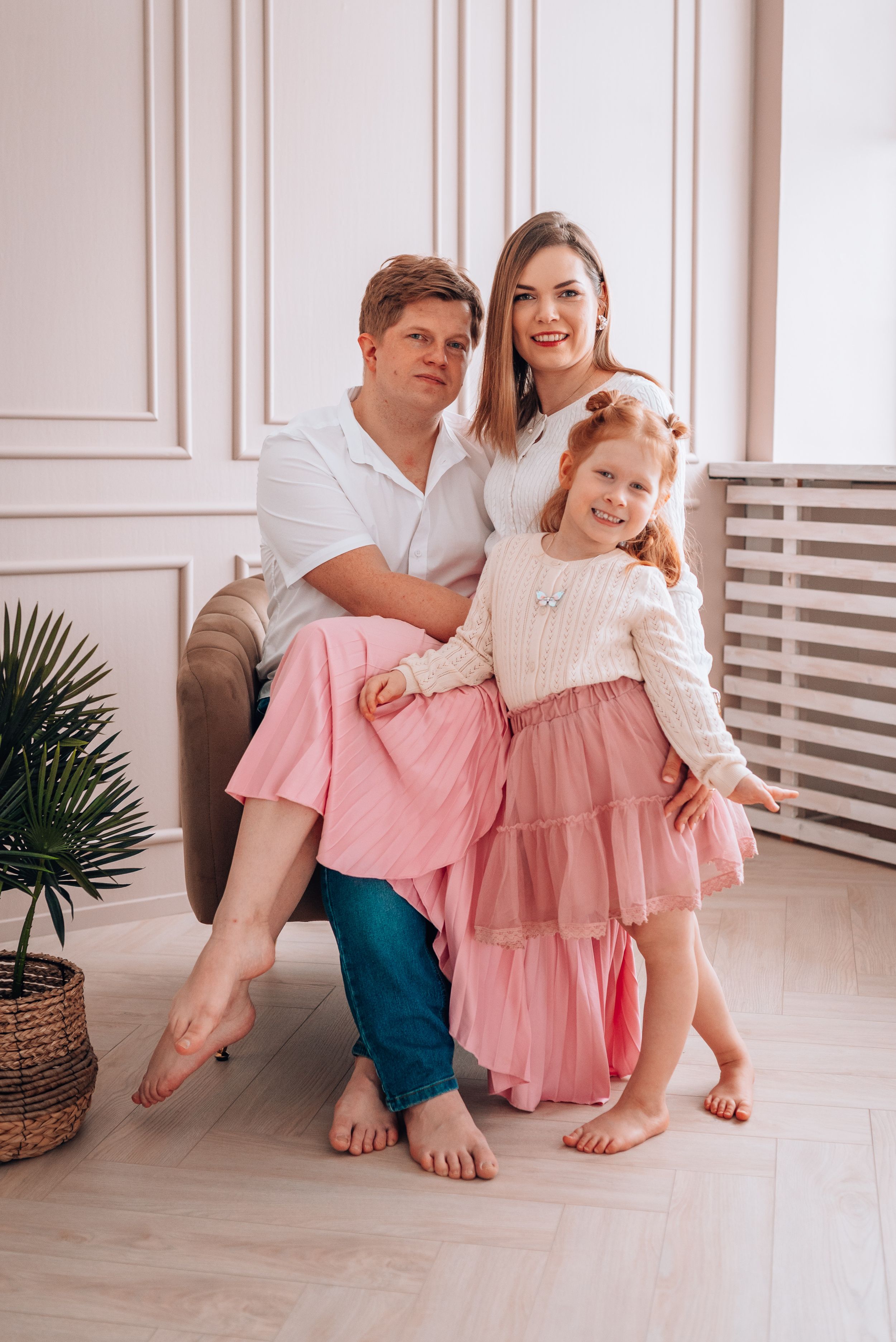 Happy family portrait with parents and redhead daughter in pink dresses posing together in a bright room.