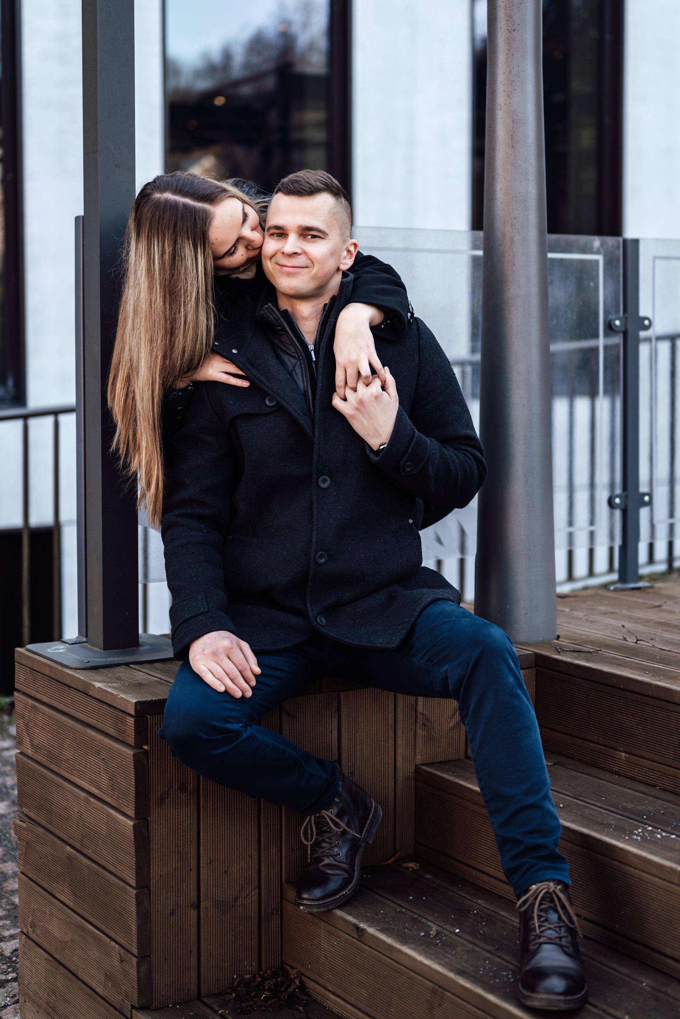 A couple sits together on wooden steps outside a building with glass windows and white trim.