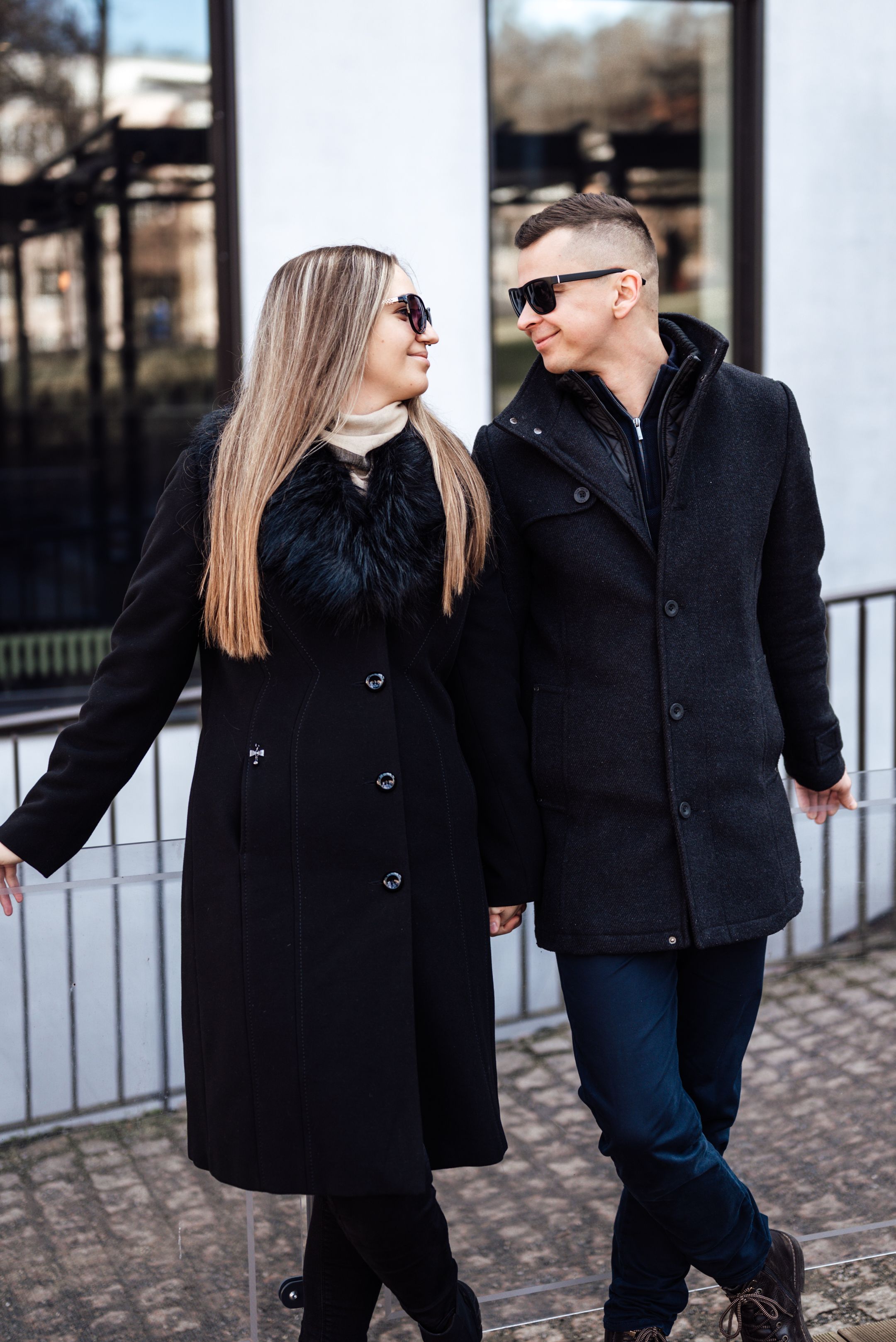 A couple in black winter coats walks together on a cobblestone path while sharing a laugh and conversation.