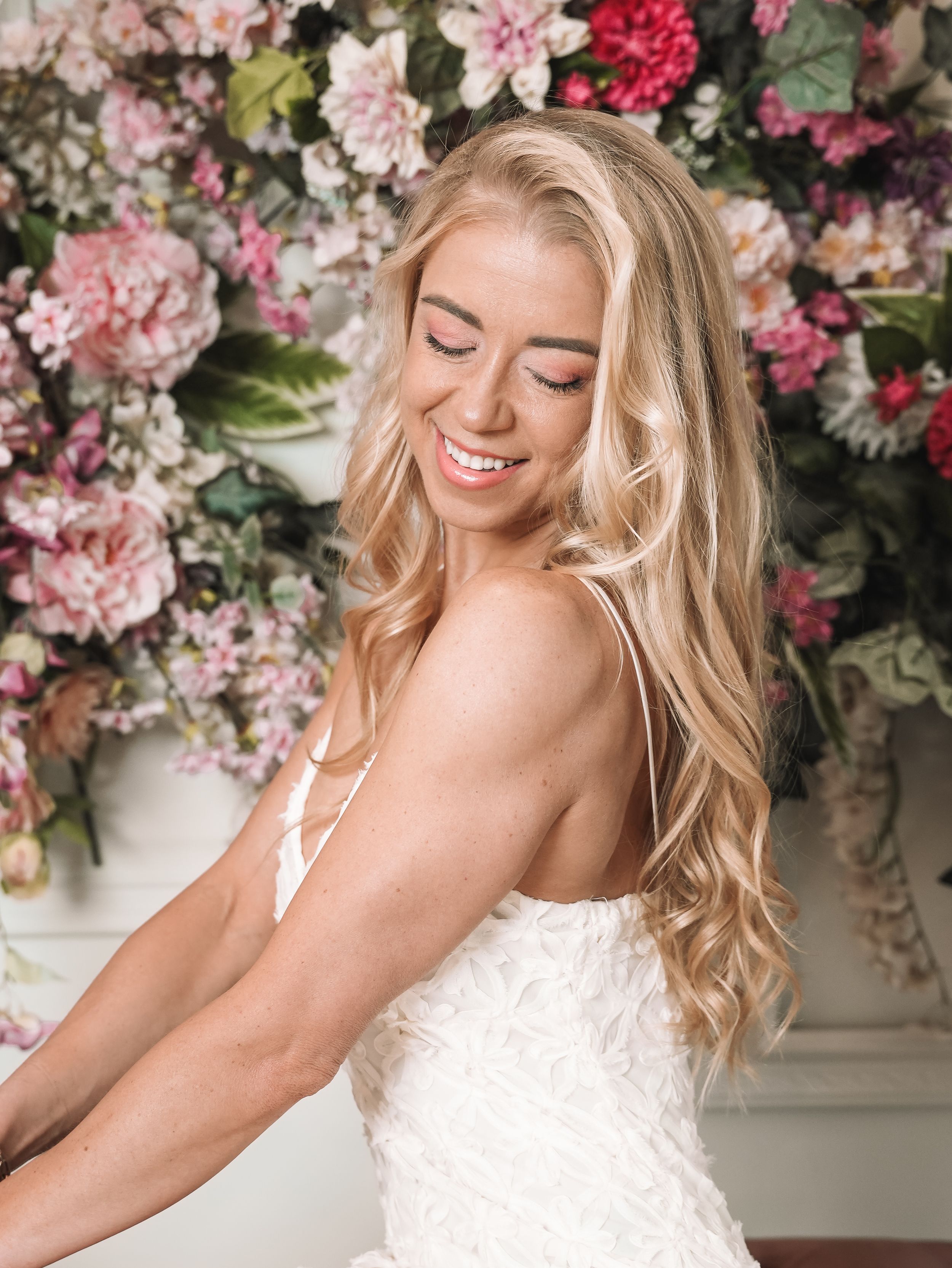 Smiling blonde bride in white lace gown poses against a lush pink floral wall backdrop.