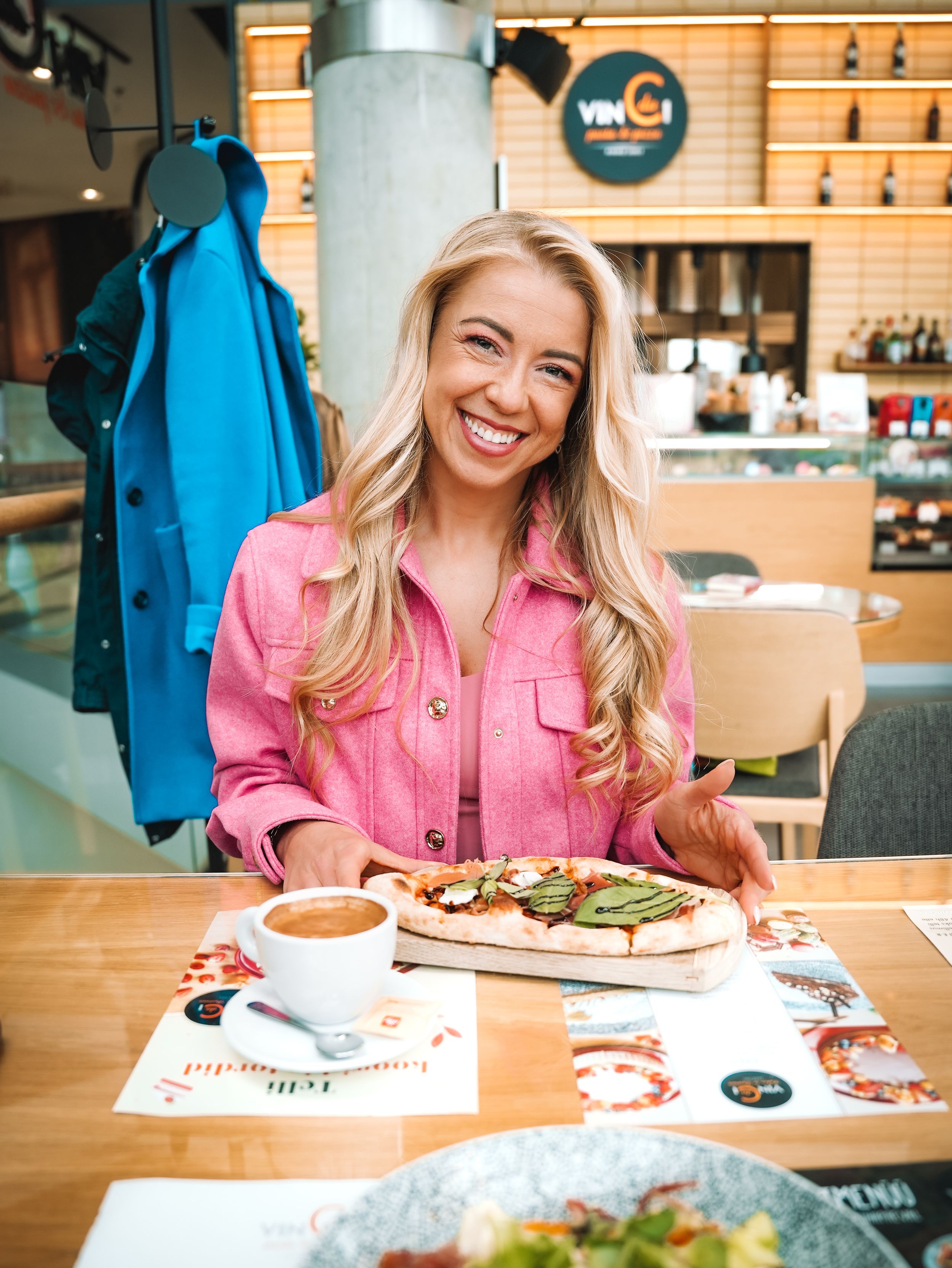 Smiling blonde woman in pink jacket enjoying pizza and coffee at a modern restaurant.