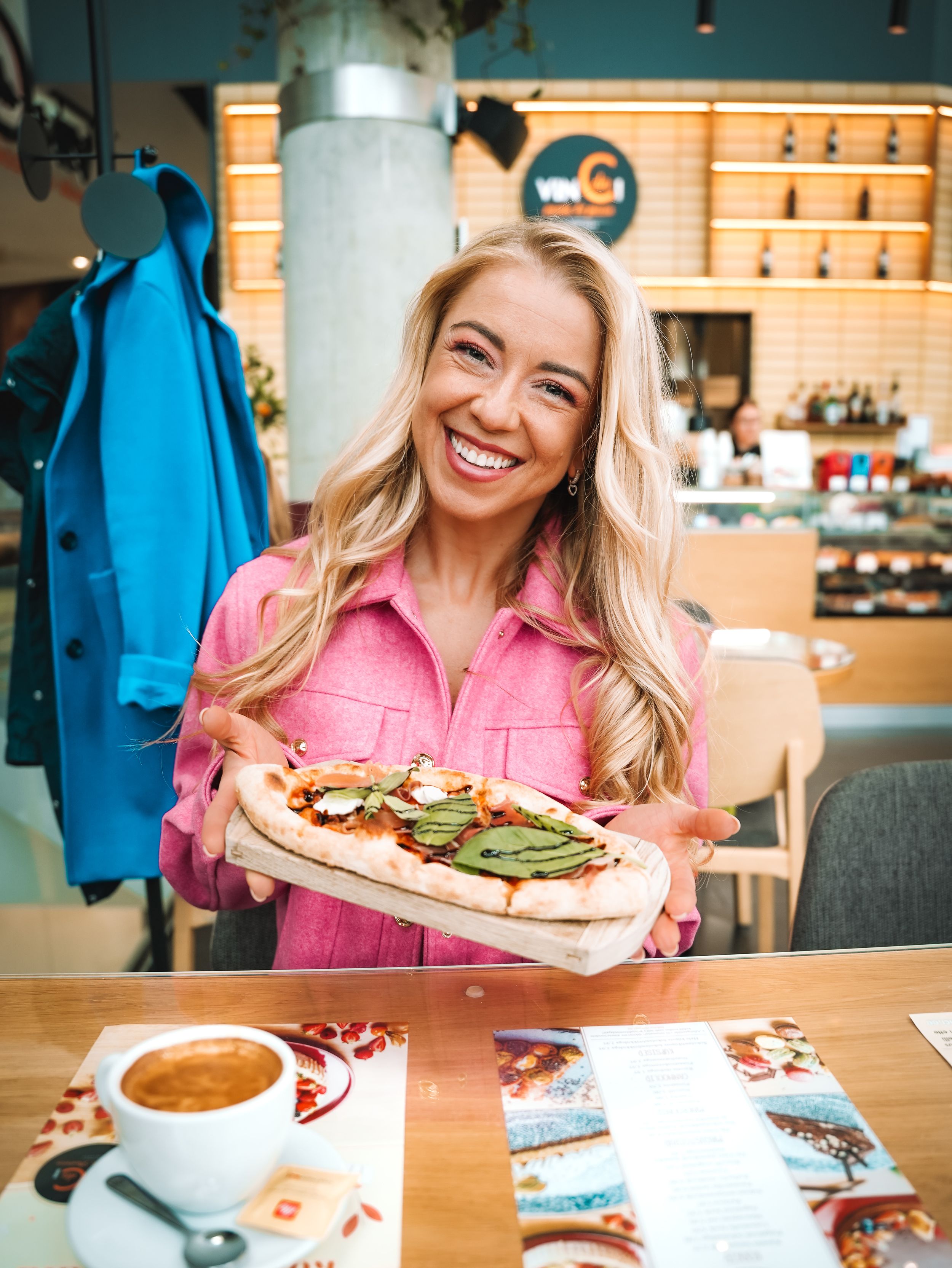 Smiling blonde woman in pink shirt holding a flatbread pizza at a modern restaurant cafe.