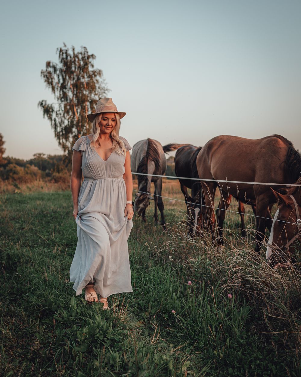 Woman in white maxi dress and hat walks near horses grazing by a fence at golden hour on a farm.